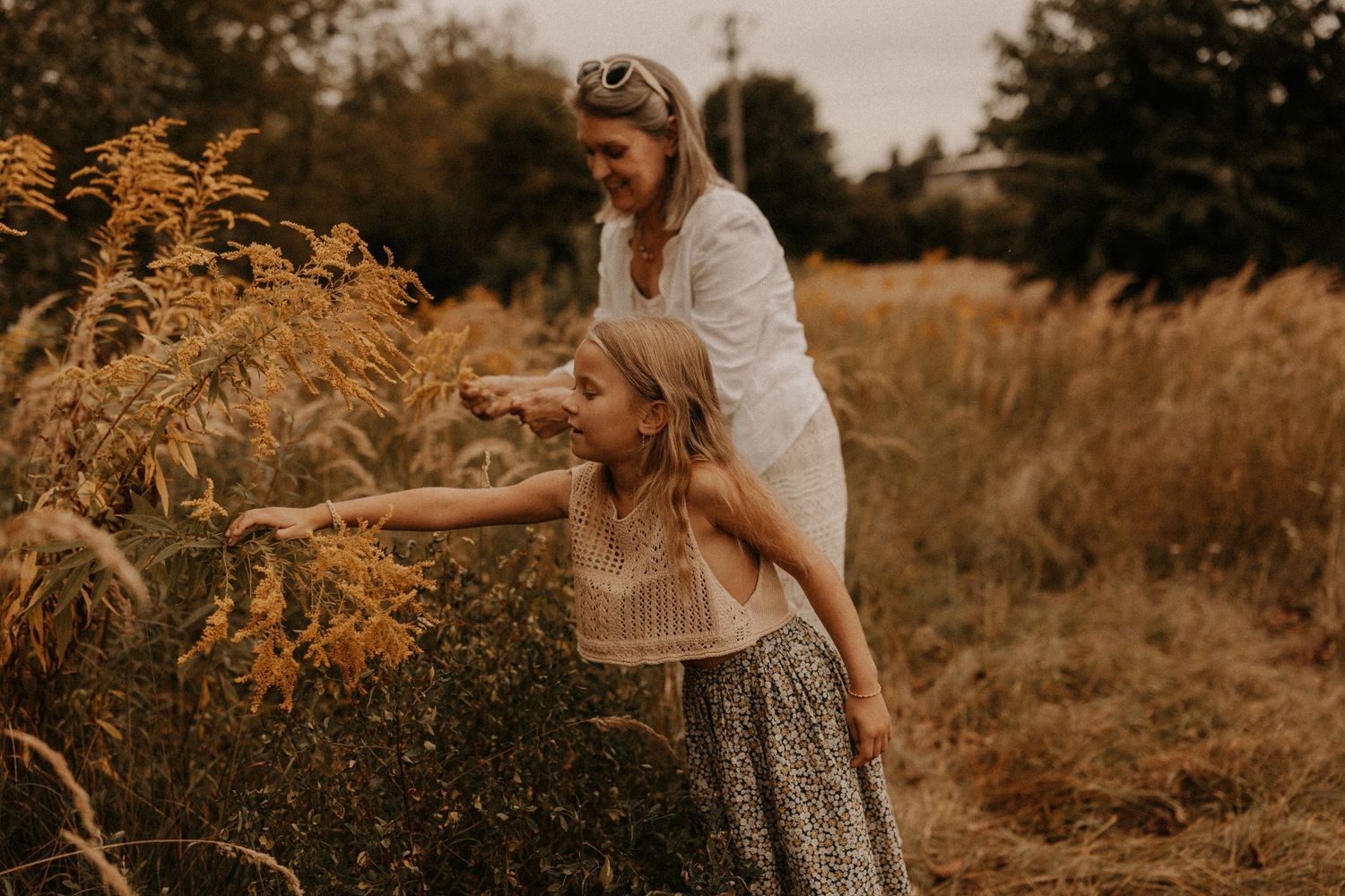 A mother and young daughter reach toward golden wildflowers in a sunlit field during a warm autumn day.