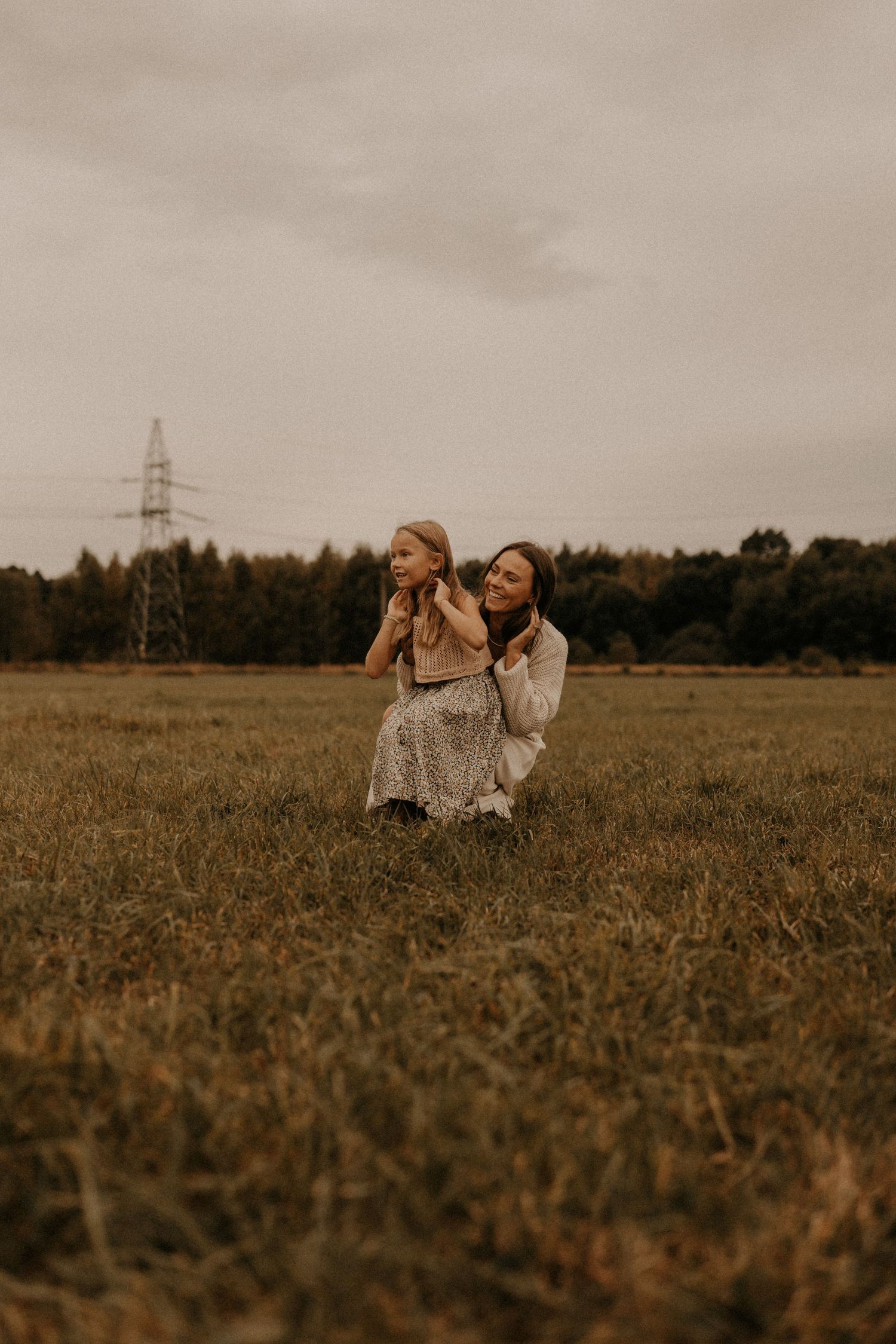 A mother kneels in a grassy field holding her young child, with trees and a power tower visible under overcast skies.