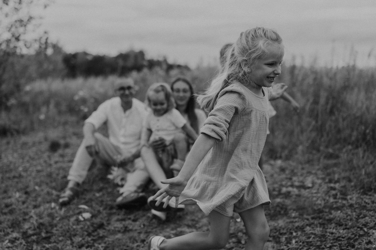 Black and white candid family photo with a young girl running in a field, family in background. sesje zdjęciowe Rybnik, Wodzisław Śląski