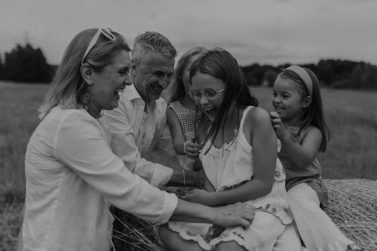 Black and white photo of a happy family laughing together on a hay bale in a field.