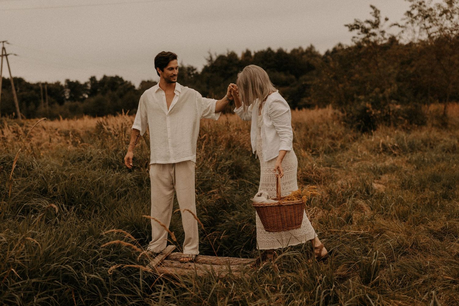 Couple in white linen outfits enjoying a romantic picnic in a golden meadow, man playfully touching woman's hair.