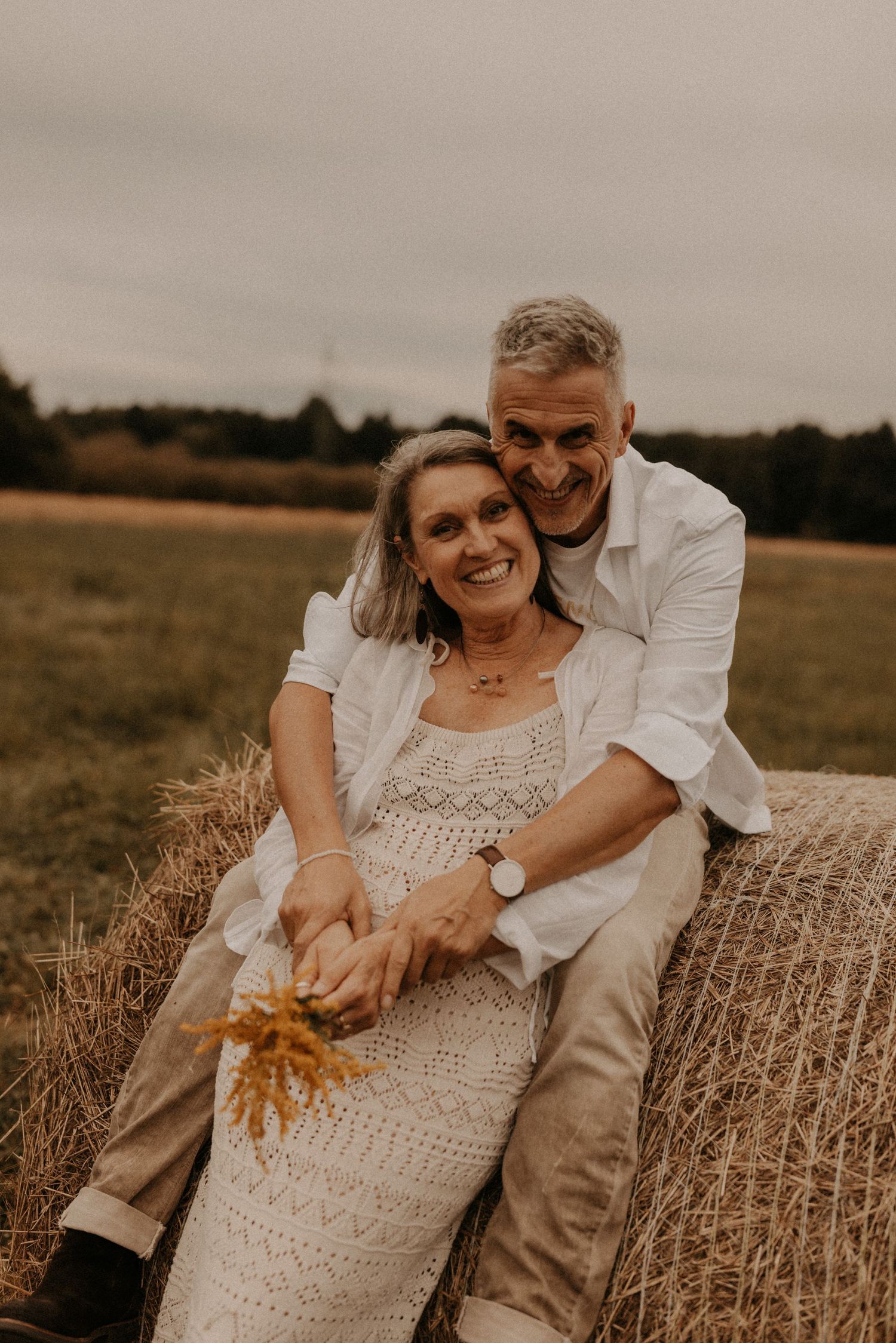 Smiling middle-aged couple sitting on a hay bale in a field, woman in white lace dress holding wildflowers.