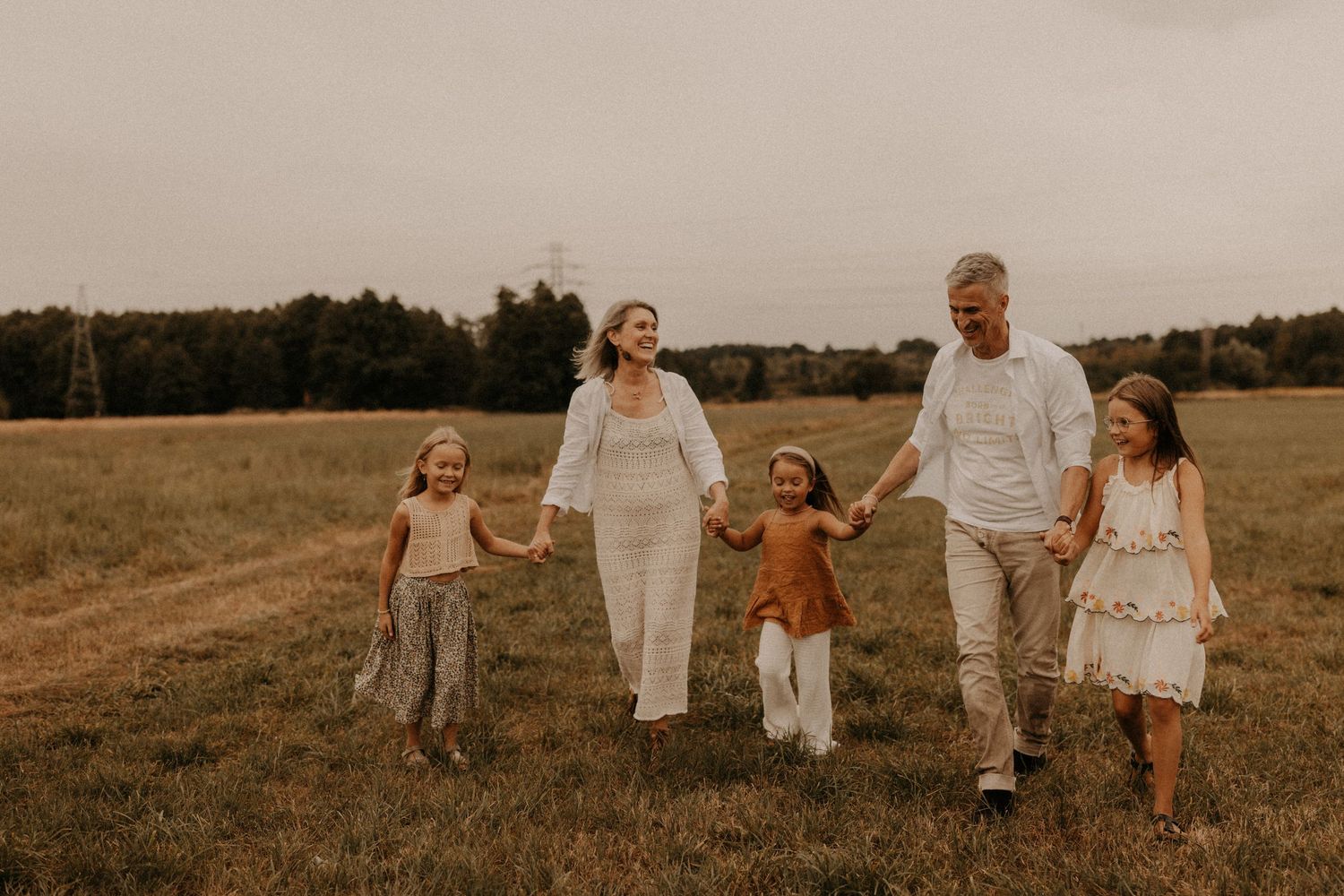 A happy family of five walks hand-in-hand through a green field, with two adults and three young girls smiling outdoors.