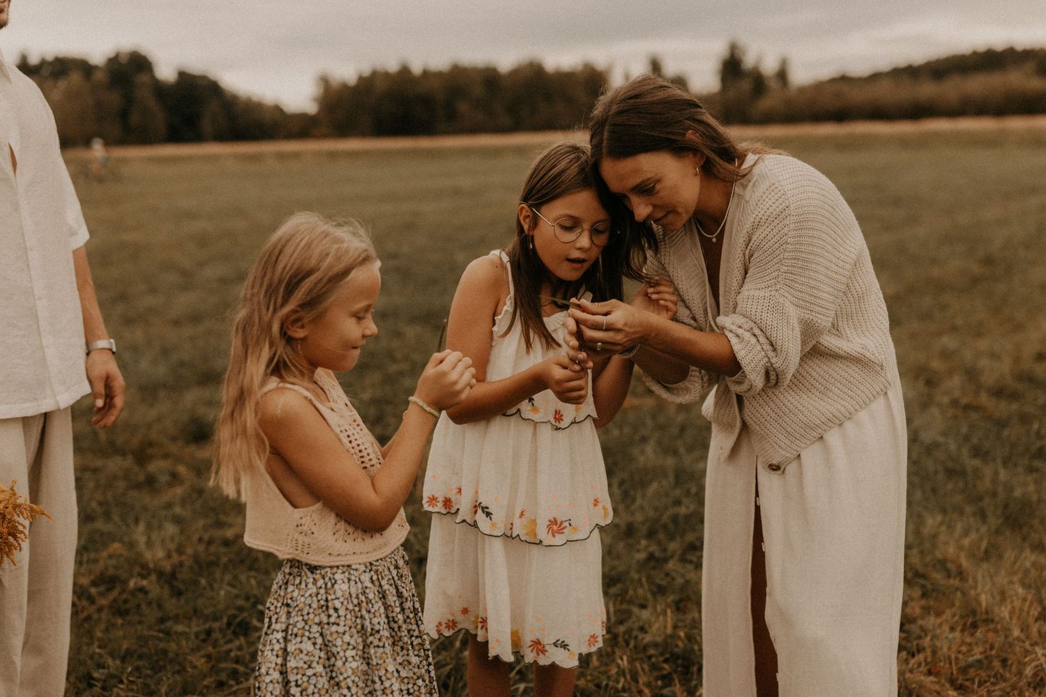 A woman in a cream cardigan shows two young girls something small in a field during a family outdoor photo session.