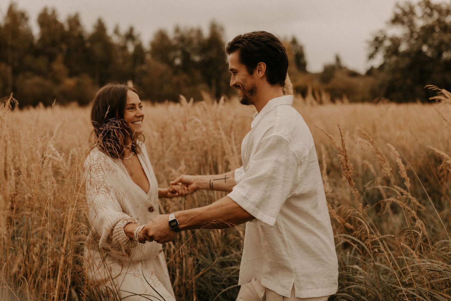 Couple holding hands and smiling in a golden wheat field, wearing neutral linen outfits in a romantic outdoor setting.