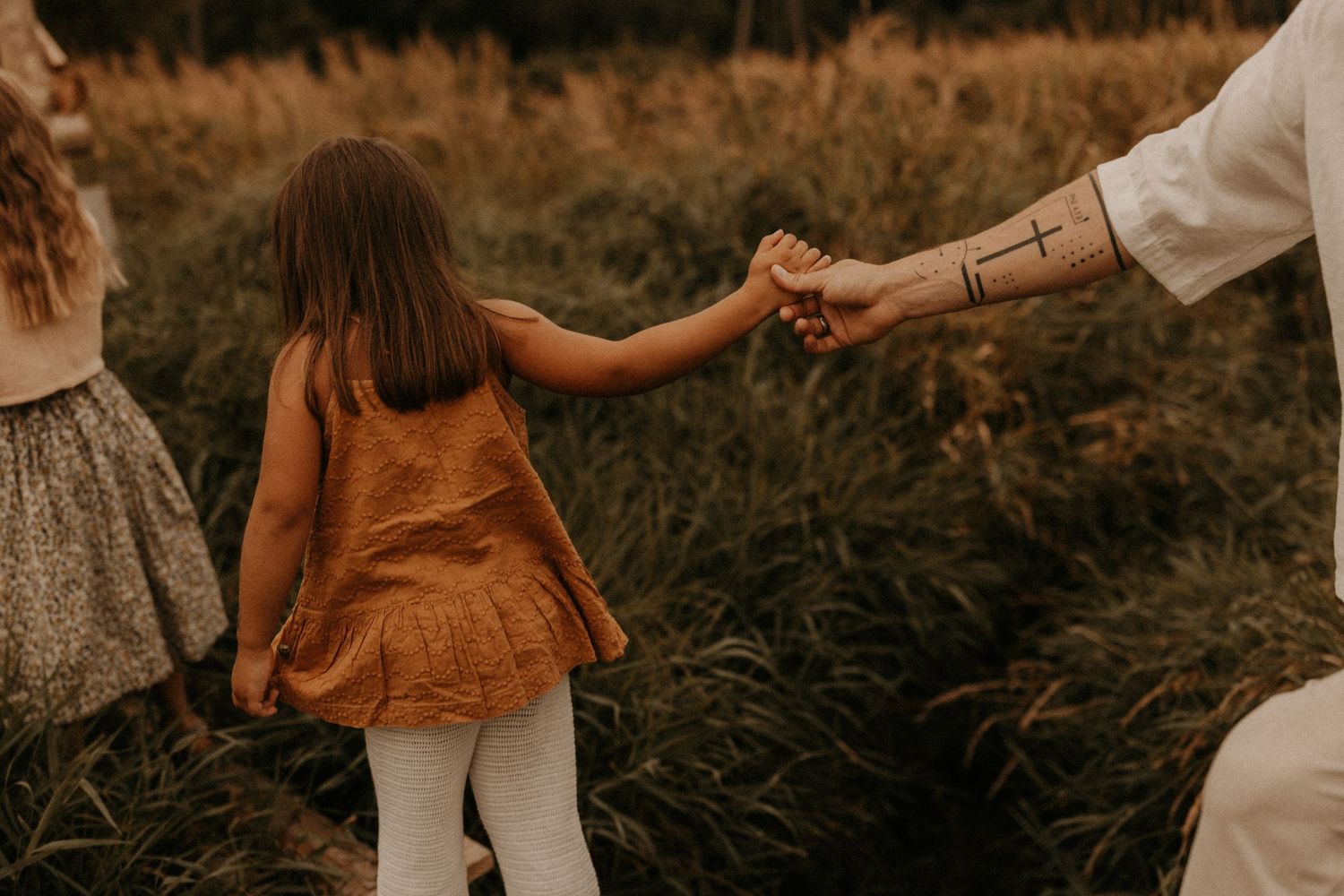 A young girl in an orange dress holds hands with a tattooed adult in a lush green field.