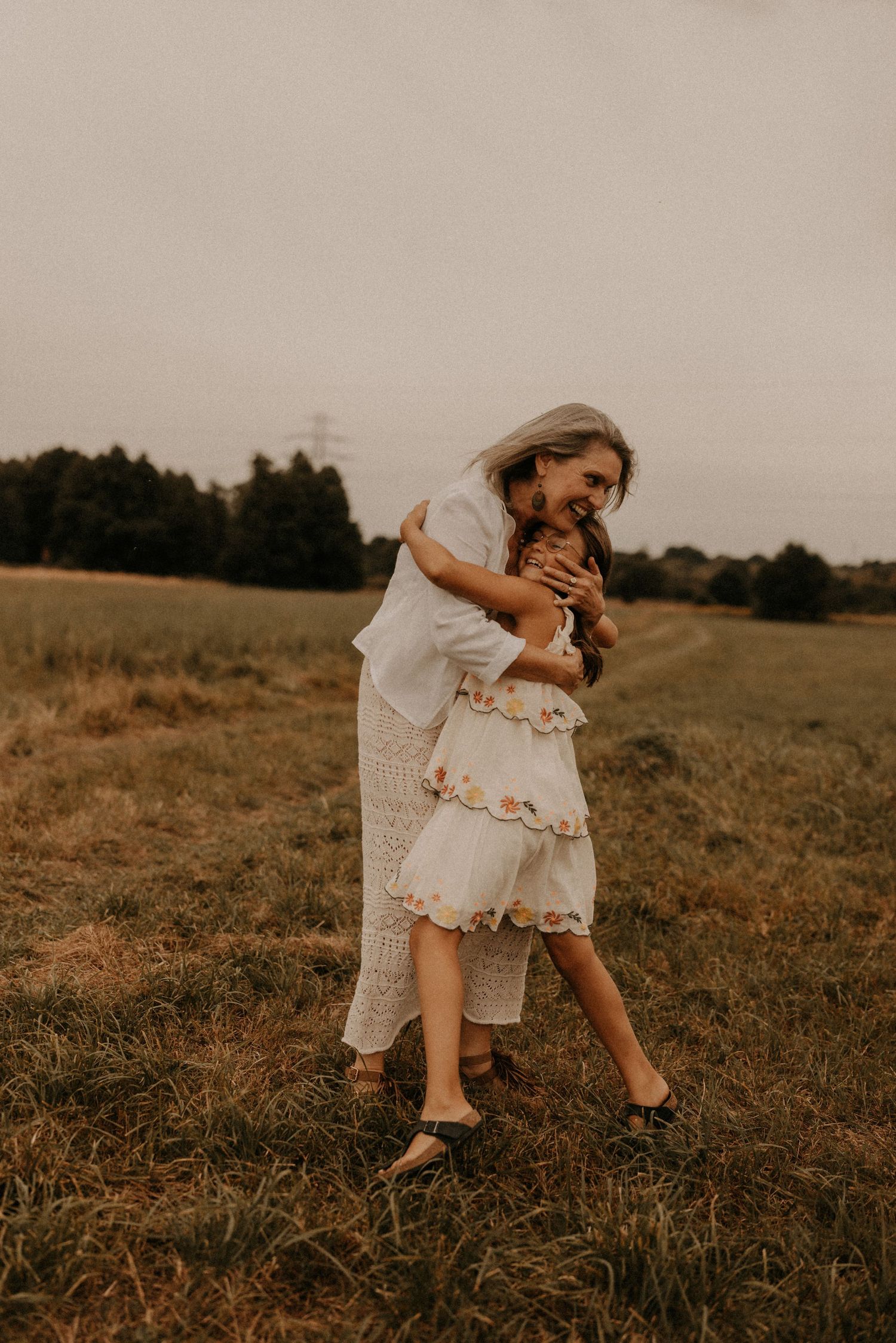 A mother and daughter share a warm embrace in a golden field, both wearing white dresses on a cloudy day.