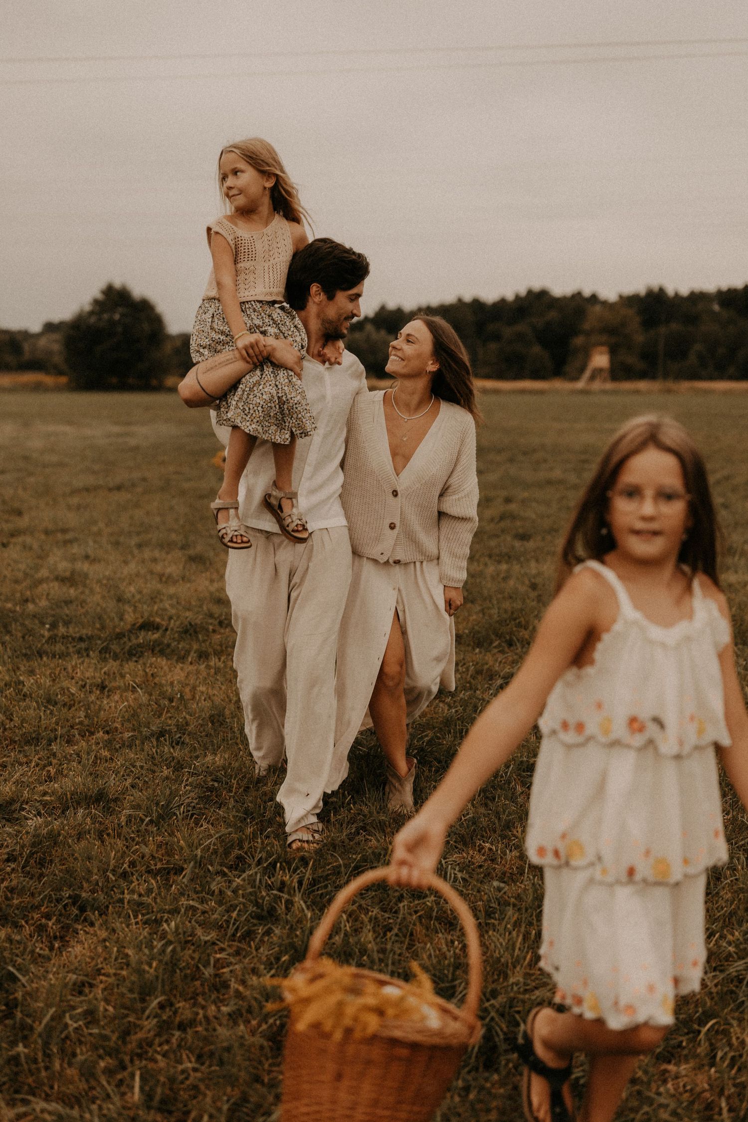 A family walks through a field, father carrying daughter on shoulders, mother smiling, girl holding picnic basket.