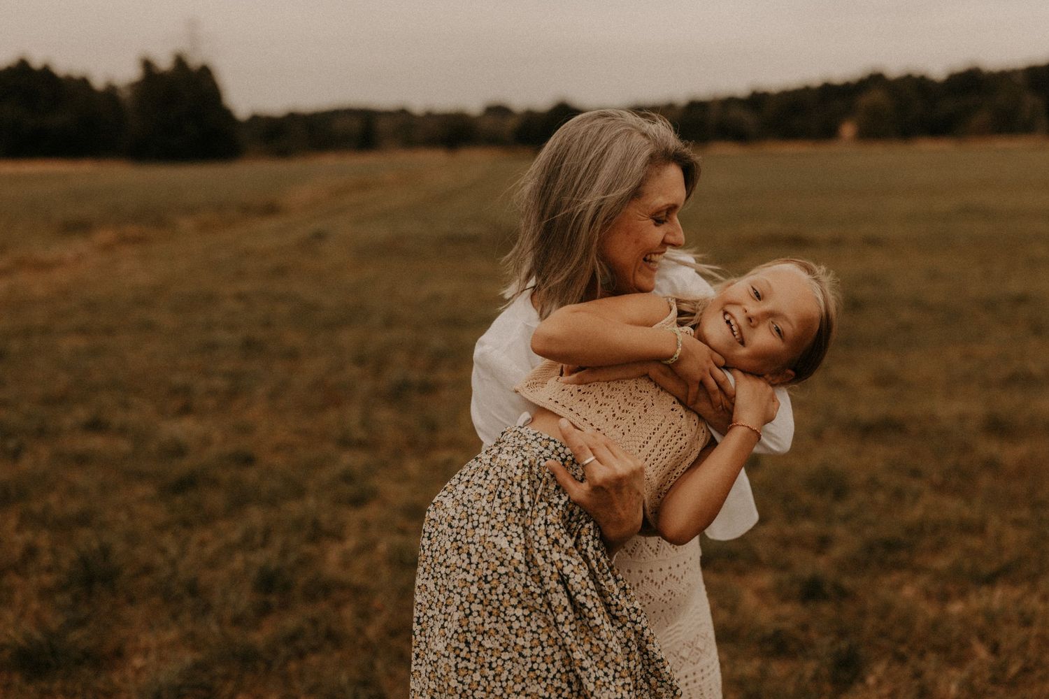 A mother joyfully embraces her young daughter in an open grassy field at dusk, both smiling warmly.