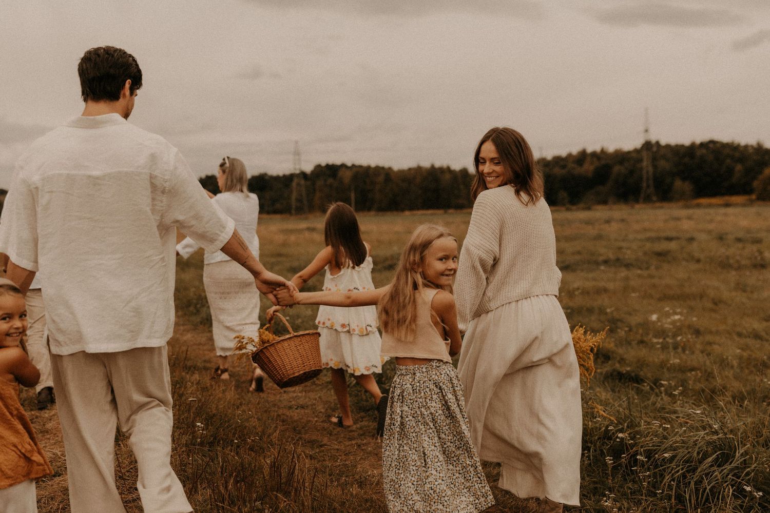 Family walking through a field holding hands, woman smiling back at camera, children in white dresses, outdoor setting.