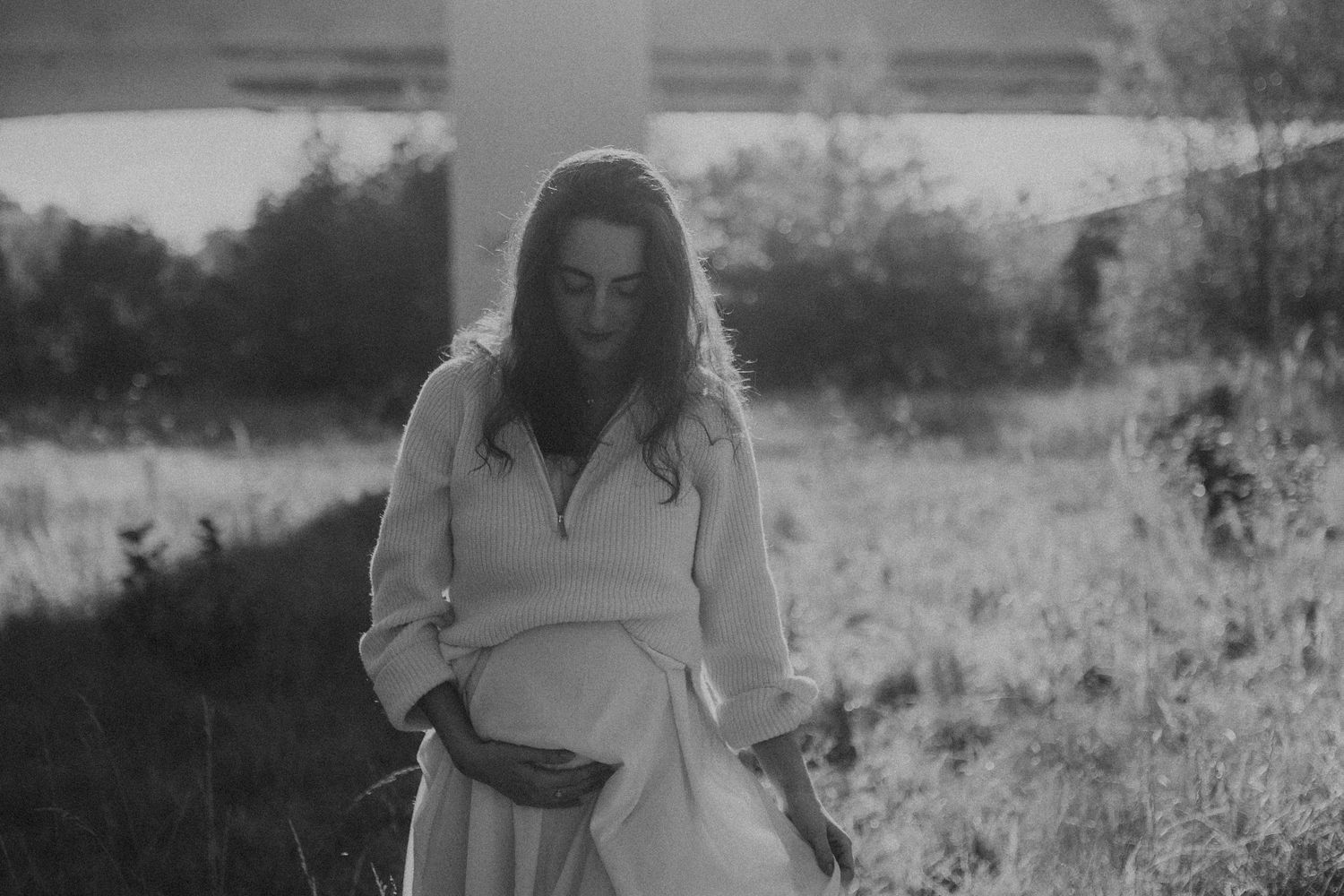 Black and white photo of a pregnant woman in a white dress cradling her belly in a field. sesje zdjęciowe Rybnik, Wodzisław Śląski