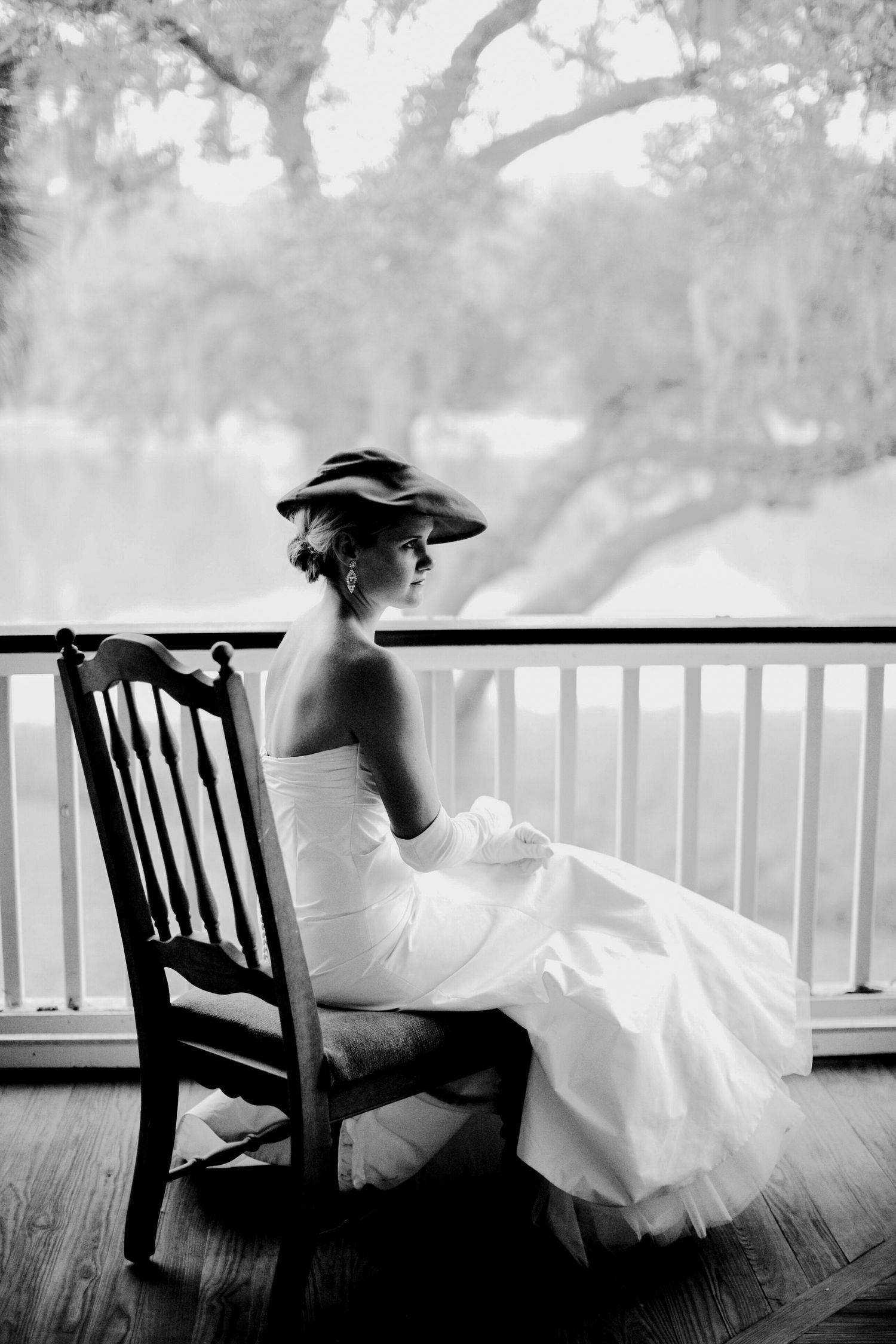 Black and white vintage style portrait of a woman in a wide-brimmed hat seated on a porch beneath a moss-draped oak