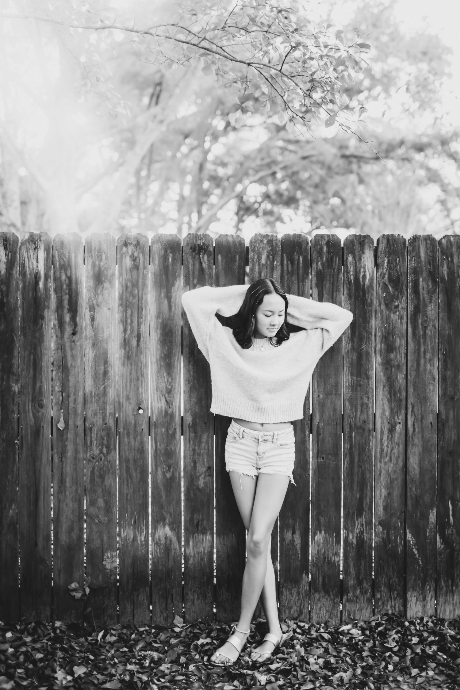 Black and white portrait of a girl in a sweater and denim shorts leaning against a weathered wooden fence with her hands behind her head