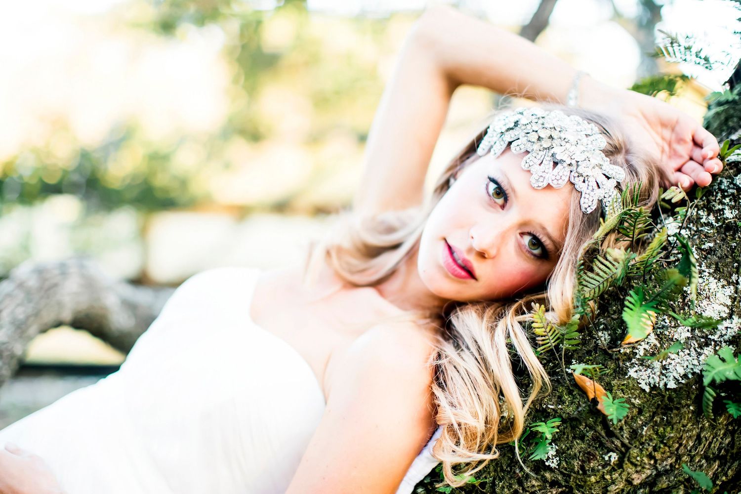 Woman wearing a jeweled headpiece reclining on a mossy tree branch in warm, glowing light