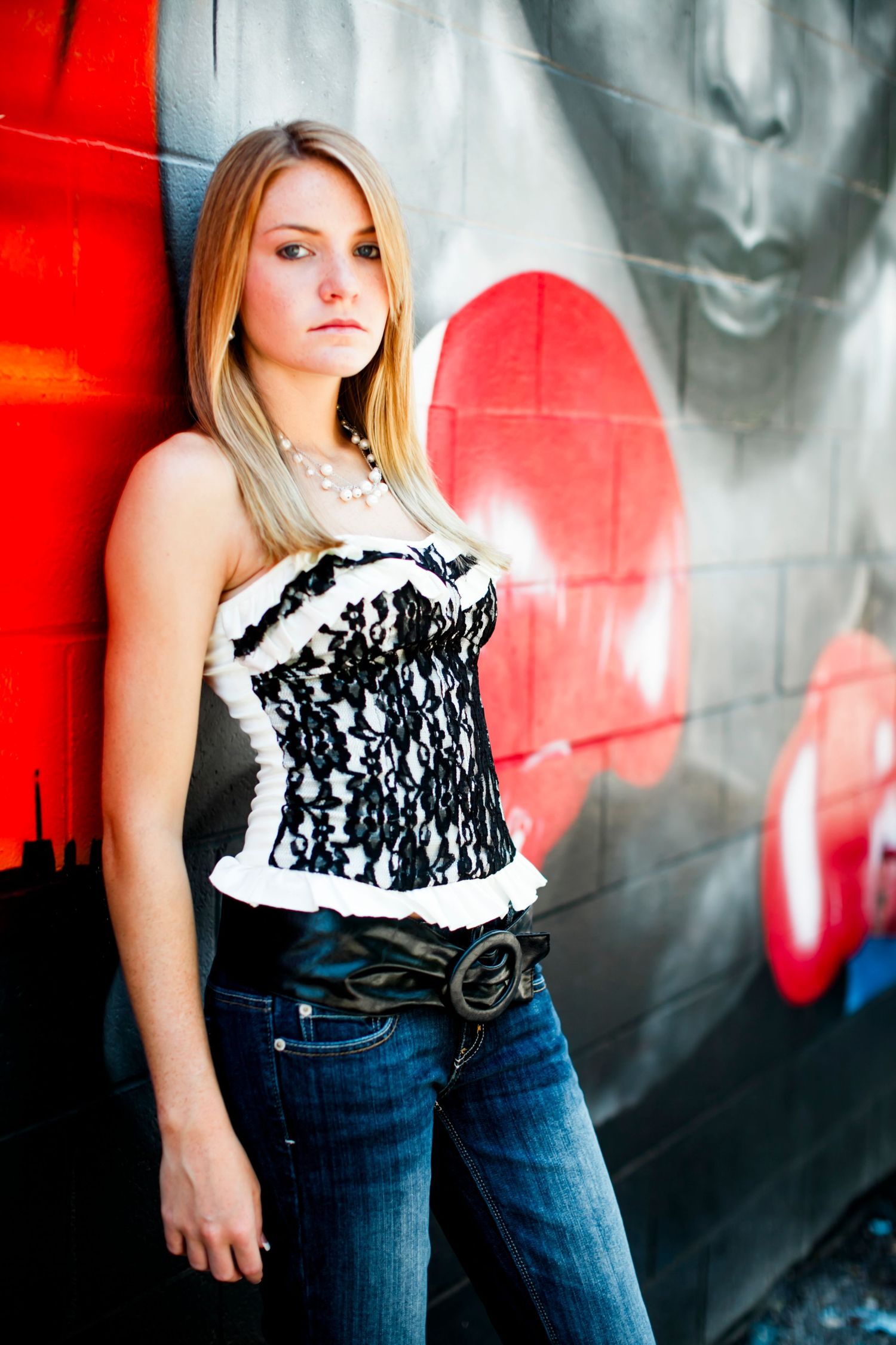 High School Senior with long blonde hair leaning against a colorful grafitti, wearing a black and white lace corset top and jeans