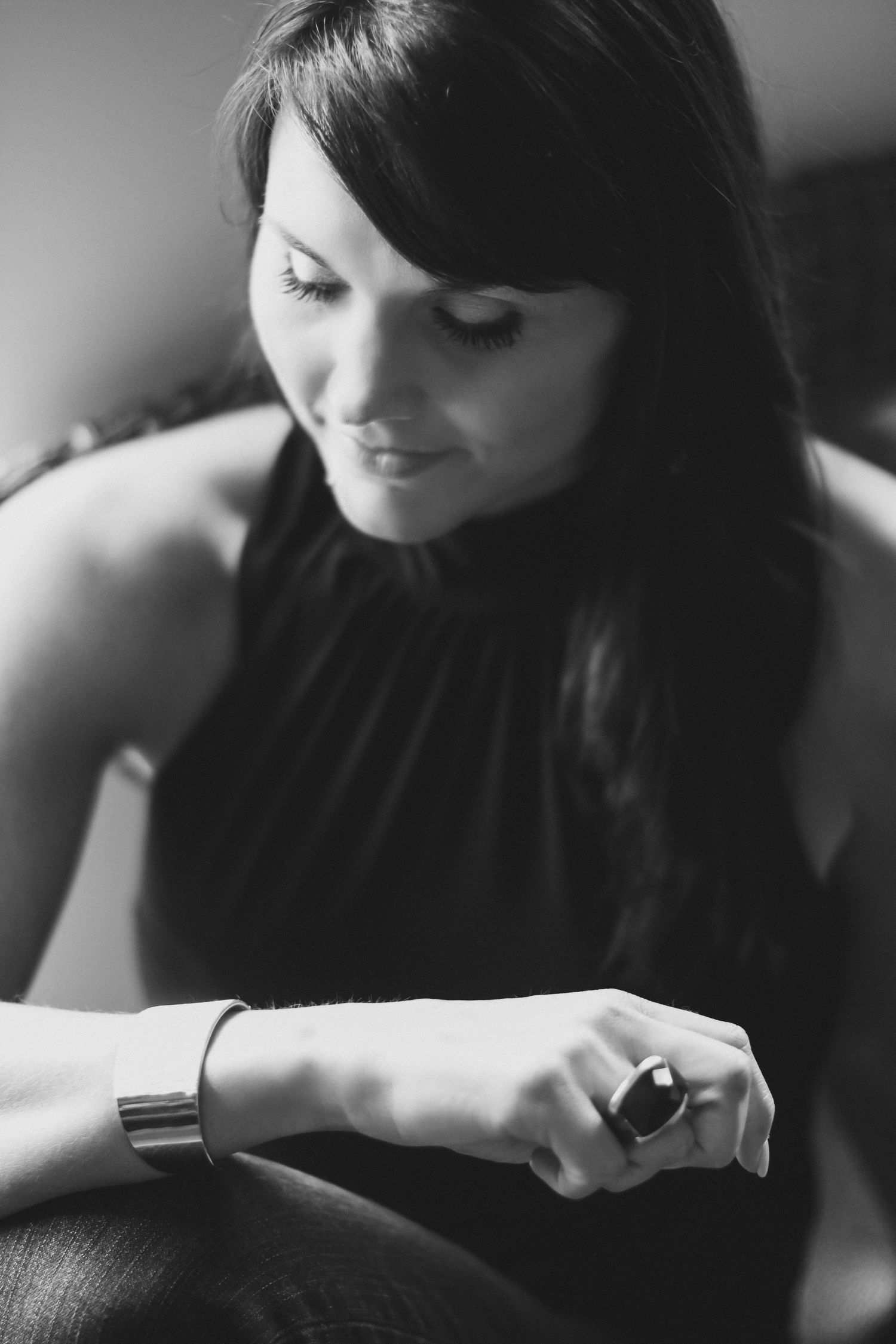 Black and white portrait of a woman with long dark hair looking down, wearing a halter top, a cuff bracelet, and a statement ring