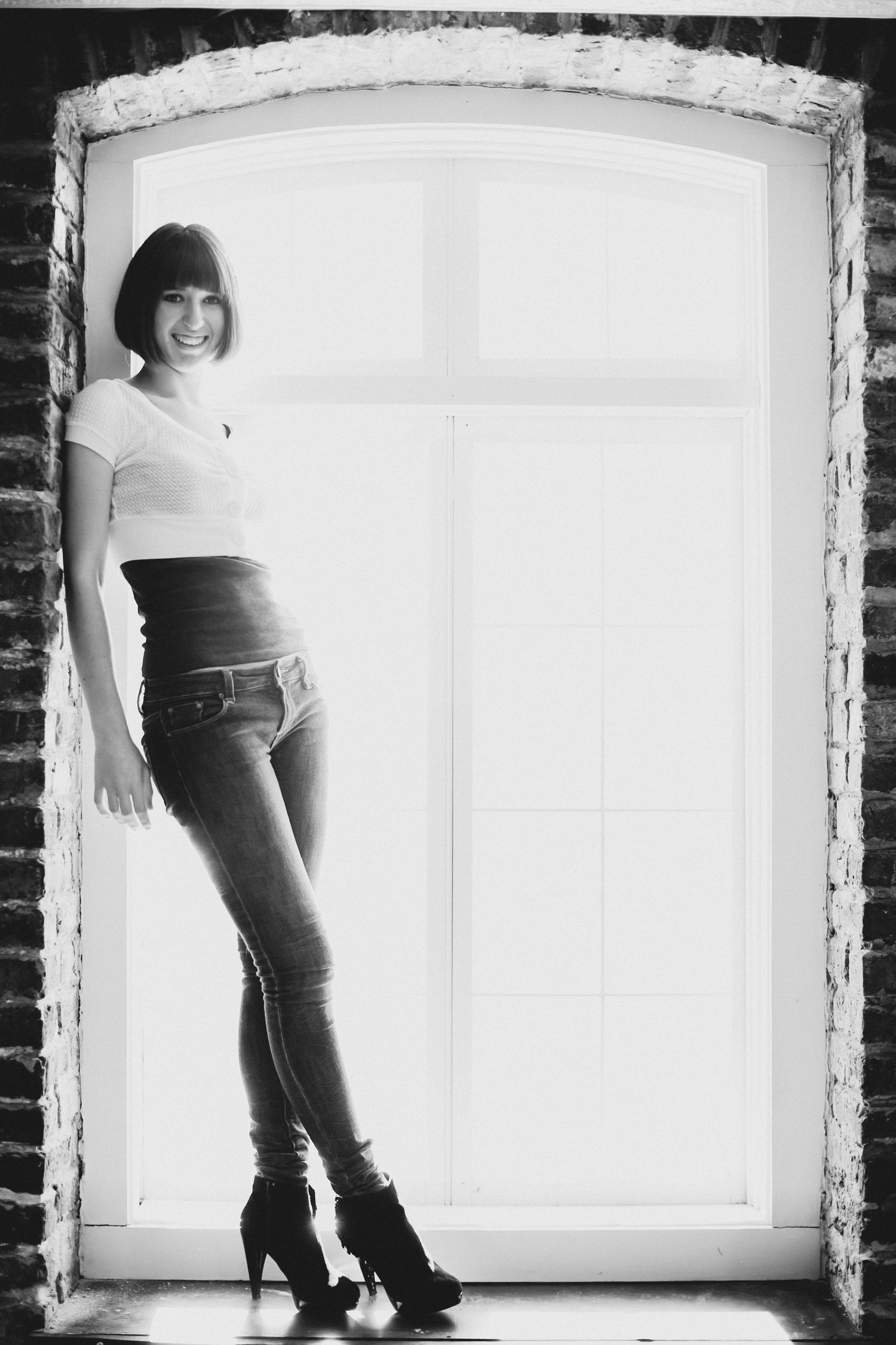 Black and white portrait of a woman in jeans and boots standing in a tall arched window frame set in old brick