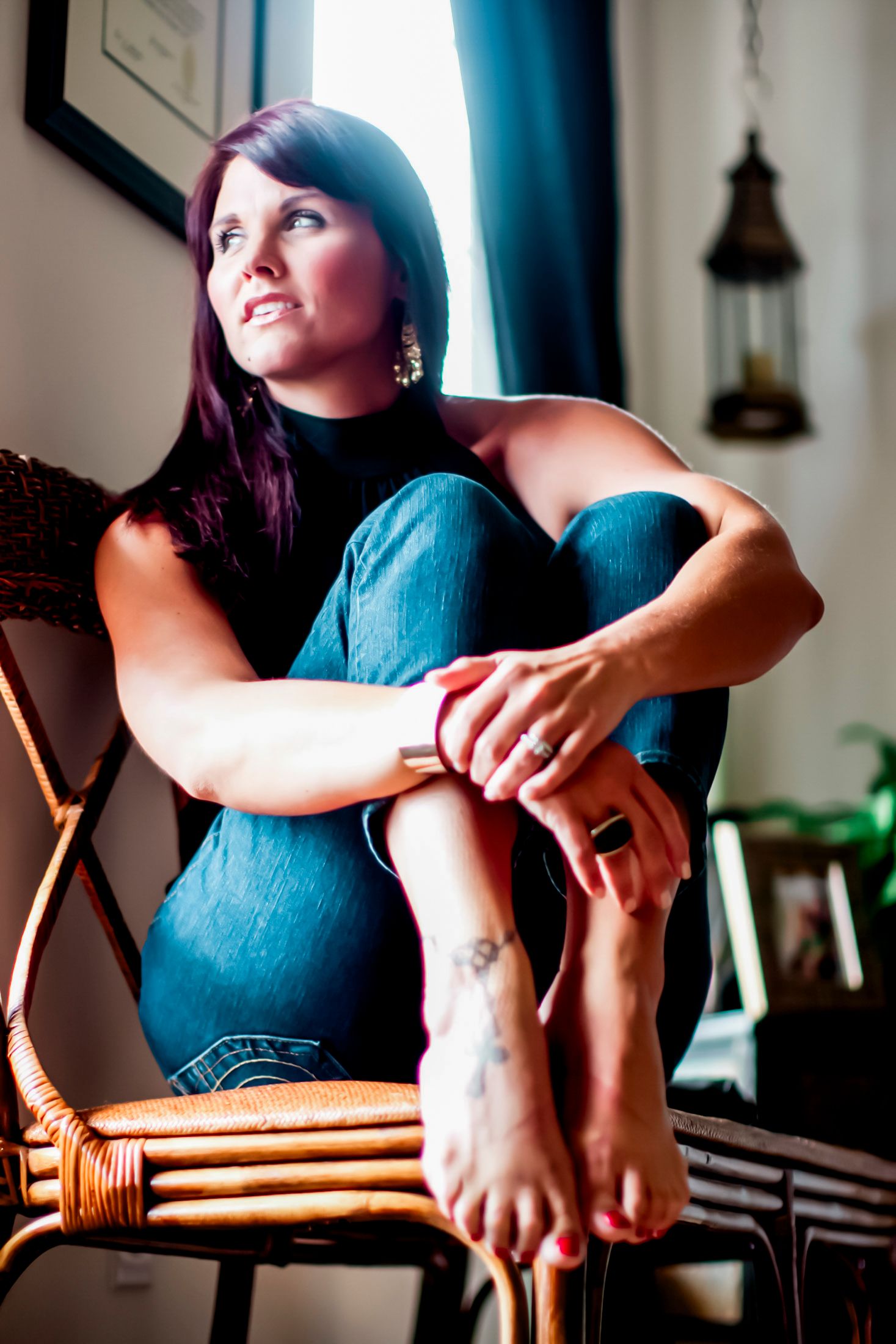 Woman with long dark hair sitting barefoot on a cane chair, gazing out a window in natural light