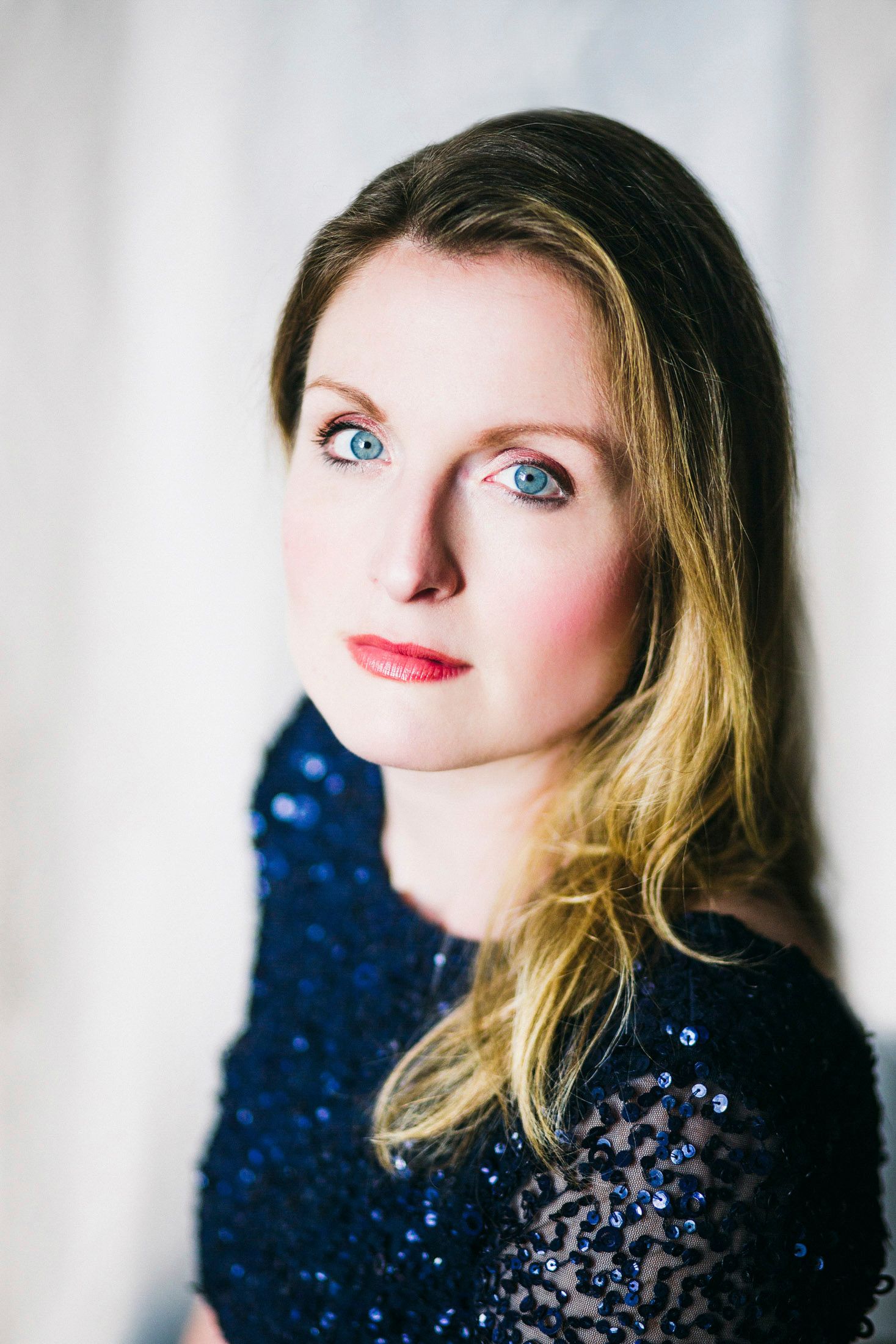 Woman with long wavy hair and blue eyes wearing a navy sequin top and red lipstick in soft window light