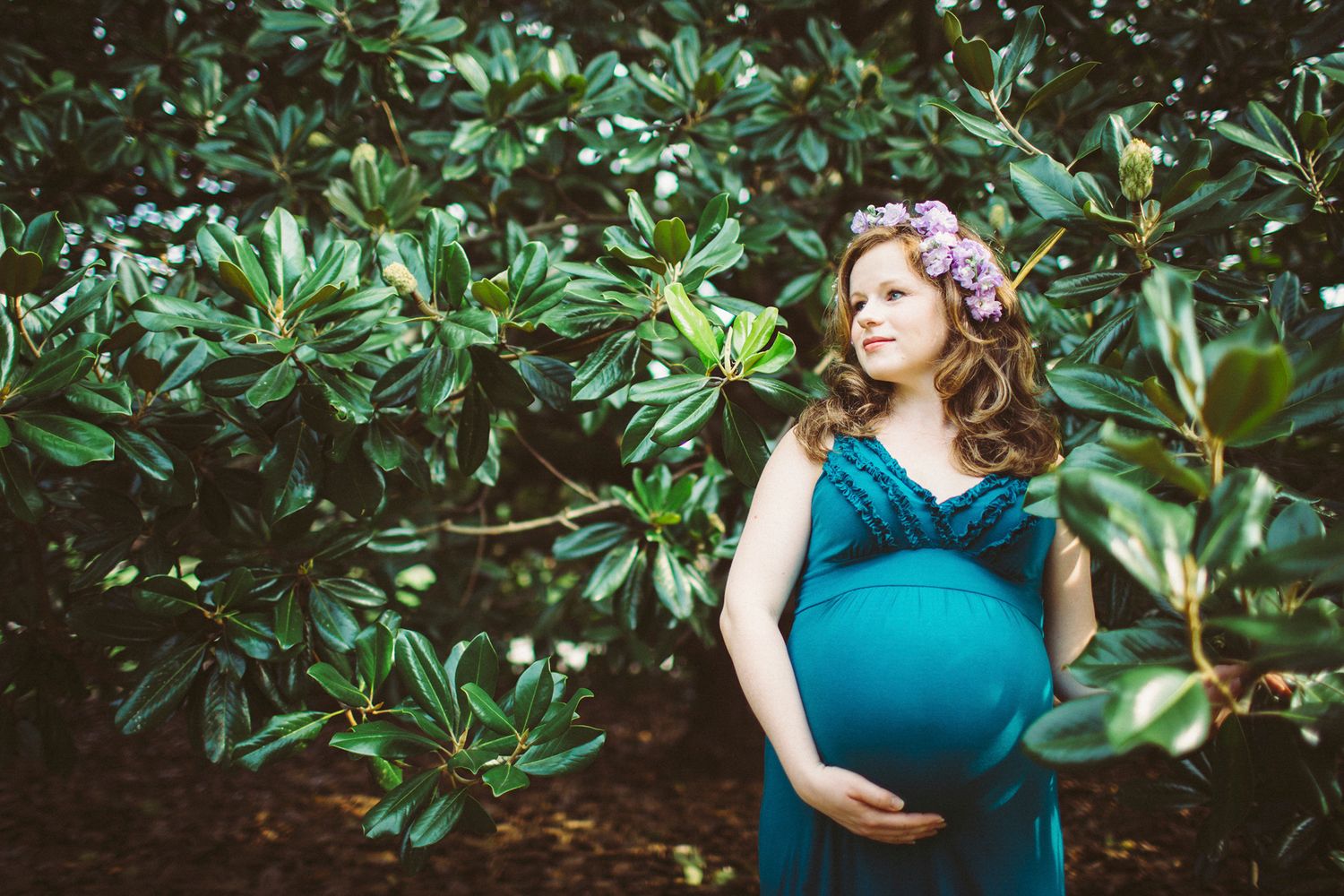 Maternity Portrait, teal dress and flower crown standing among glossy green leaves, hands resting on her belly, Hampton Park Charleston