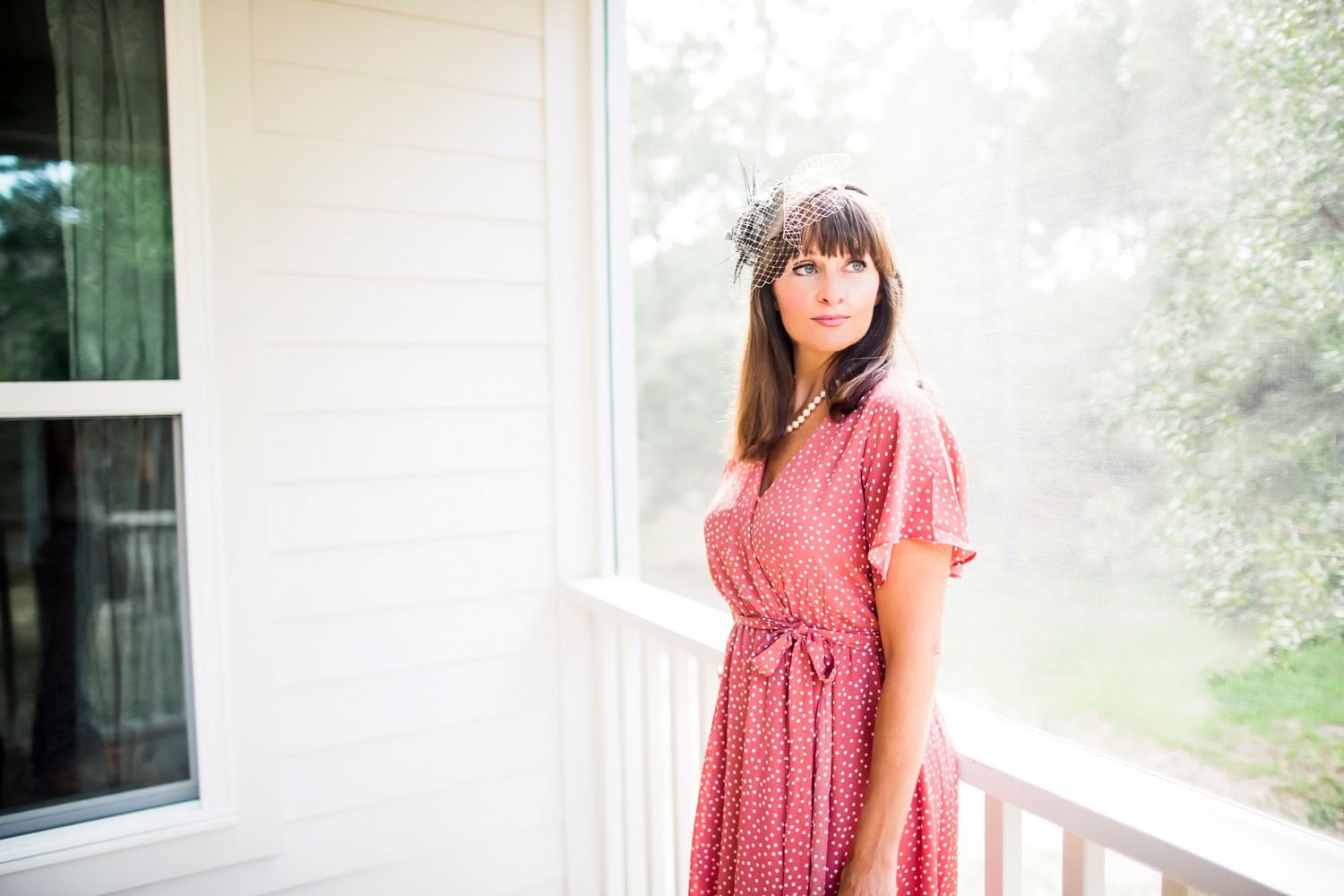 Woman in a coral-pink dress standing on a white porch in soft, hazy morning light, vintage inspired portrait