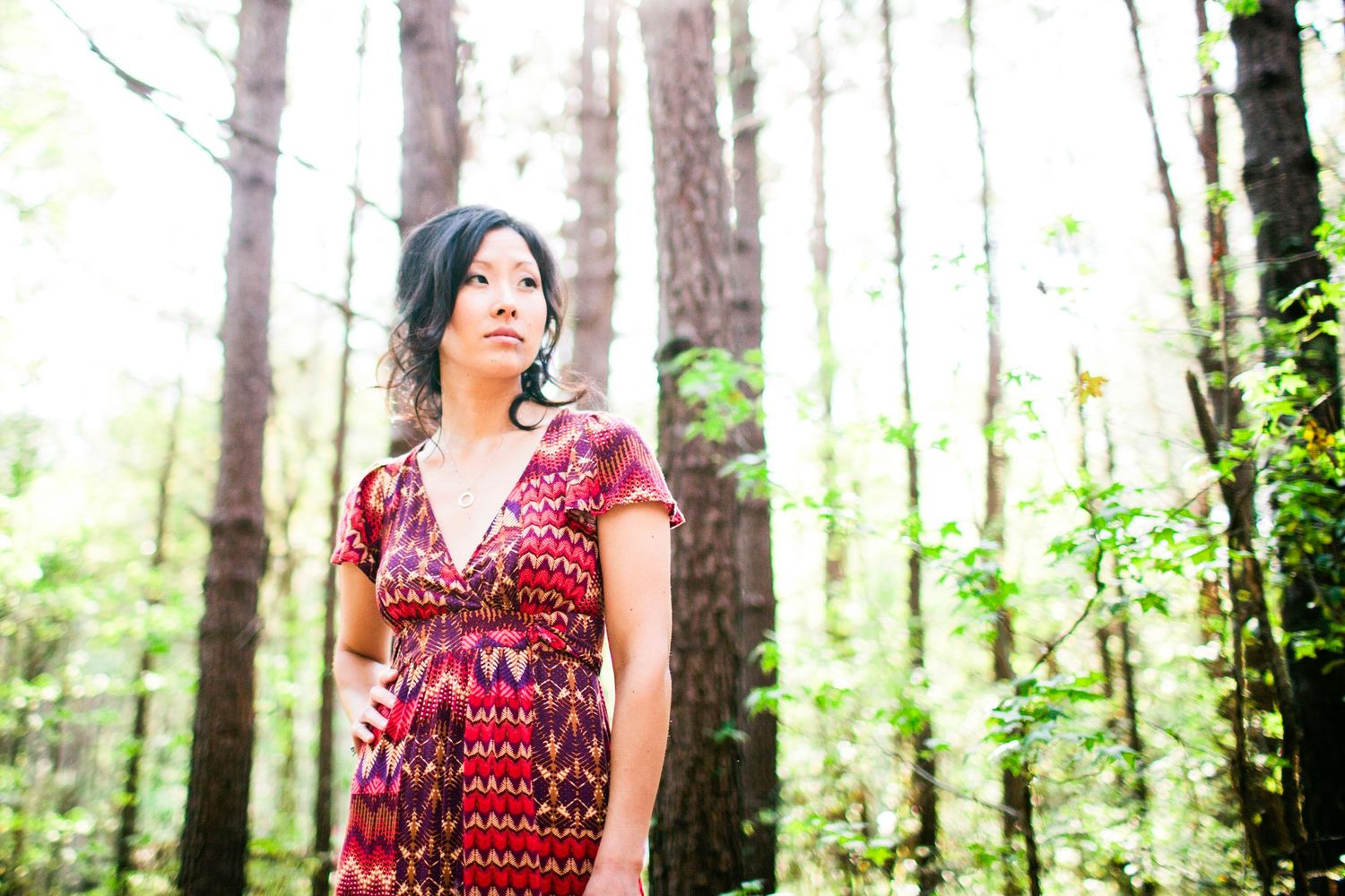 Woman in a red patterned dress standing among tall pine trees in soft natural light