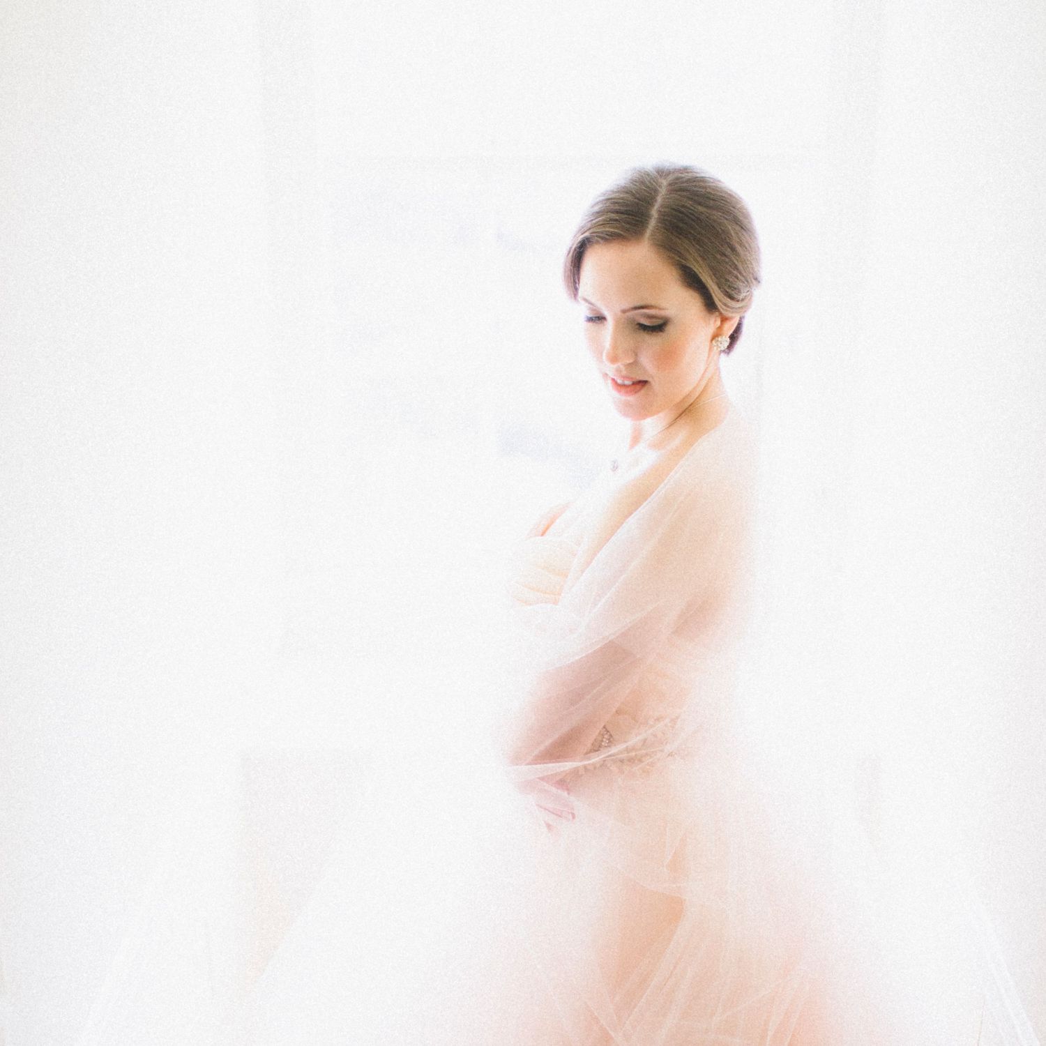 Bright, airy portrait of a bride looking down, wrapped in sheer, soft pink tulle in window light