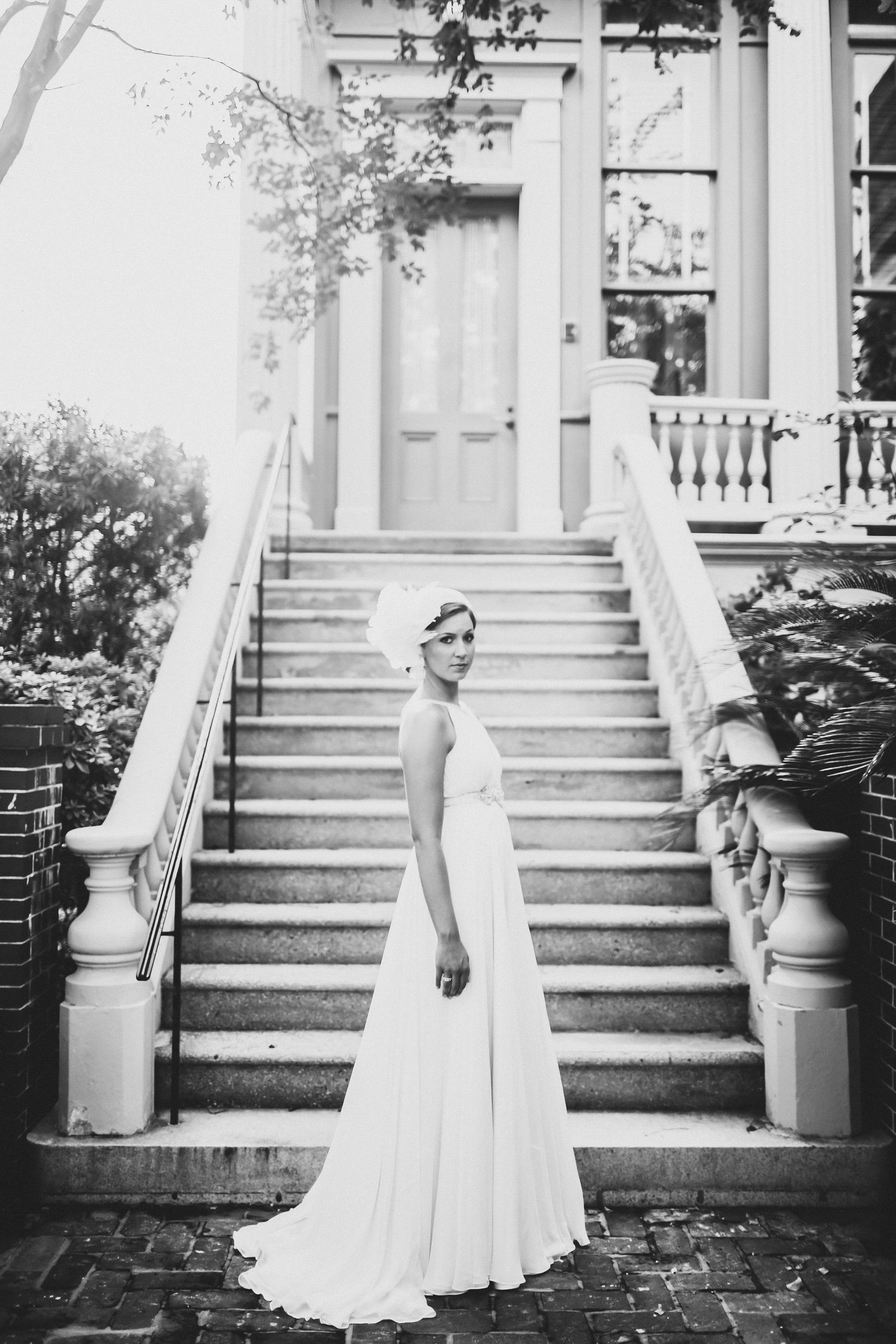 Black and white portrait of a bride in a flowing gown on a grand stone staircase in front of a columned historic home