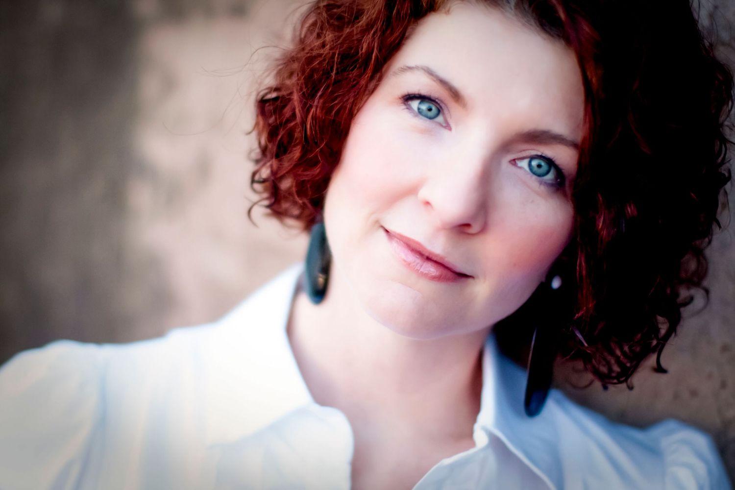 Close-up portrait of a woman with curly auburn hair and blue eyes wearing a white collared shirt and dark earrings
