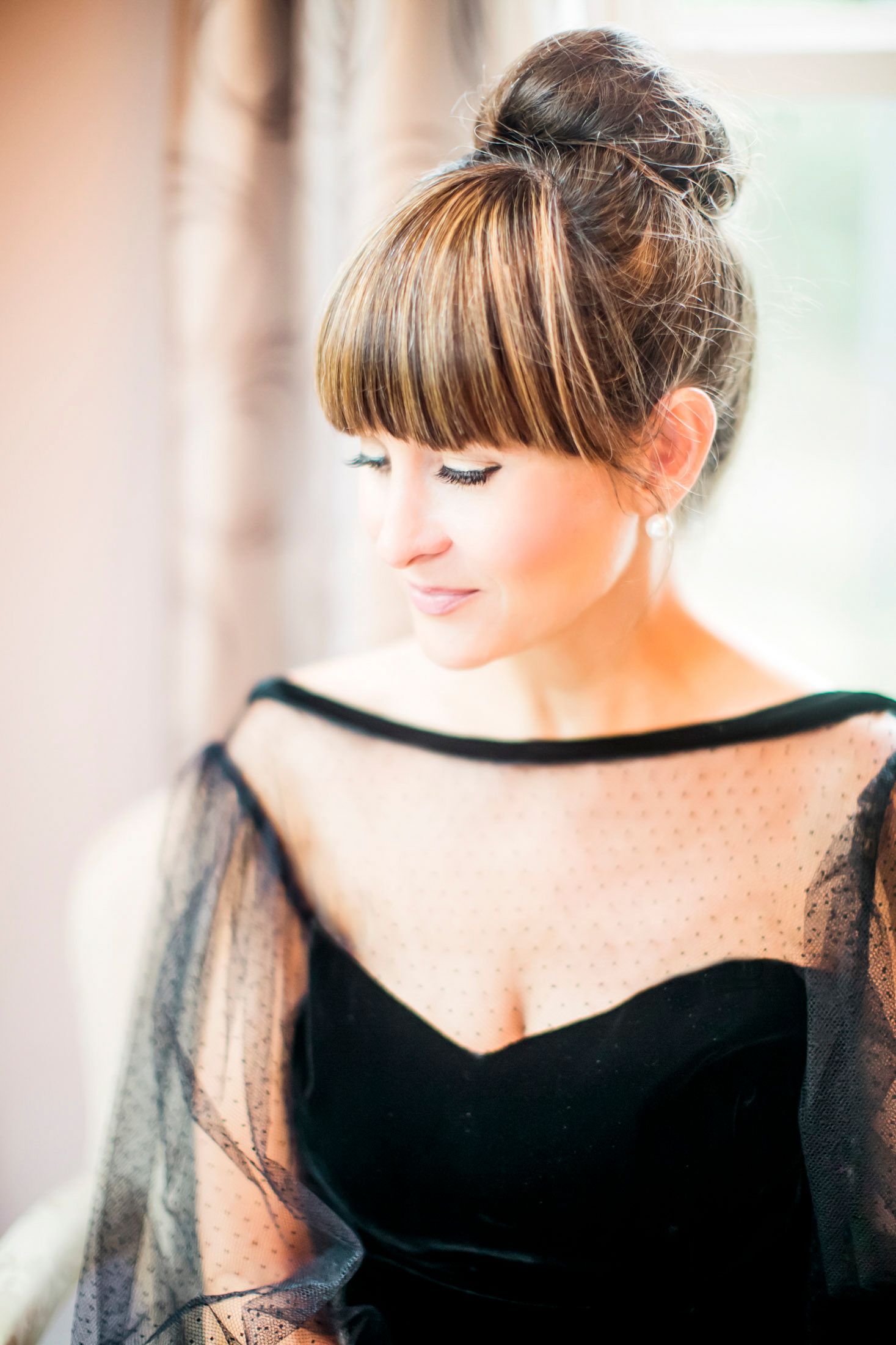 Woman with a high bun and bangs gazing softly to the side in window light, wearing a black dress with sheer sleeves