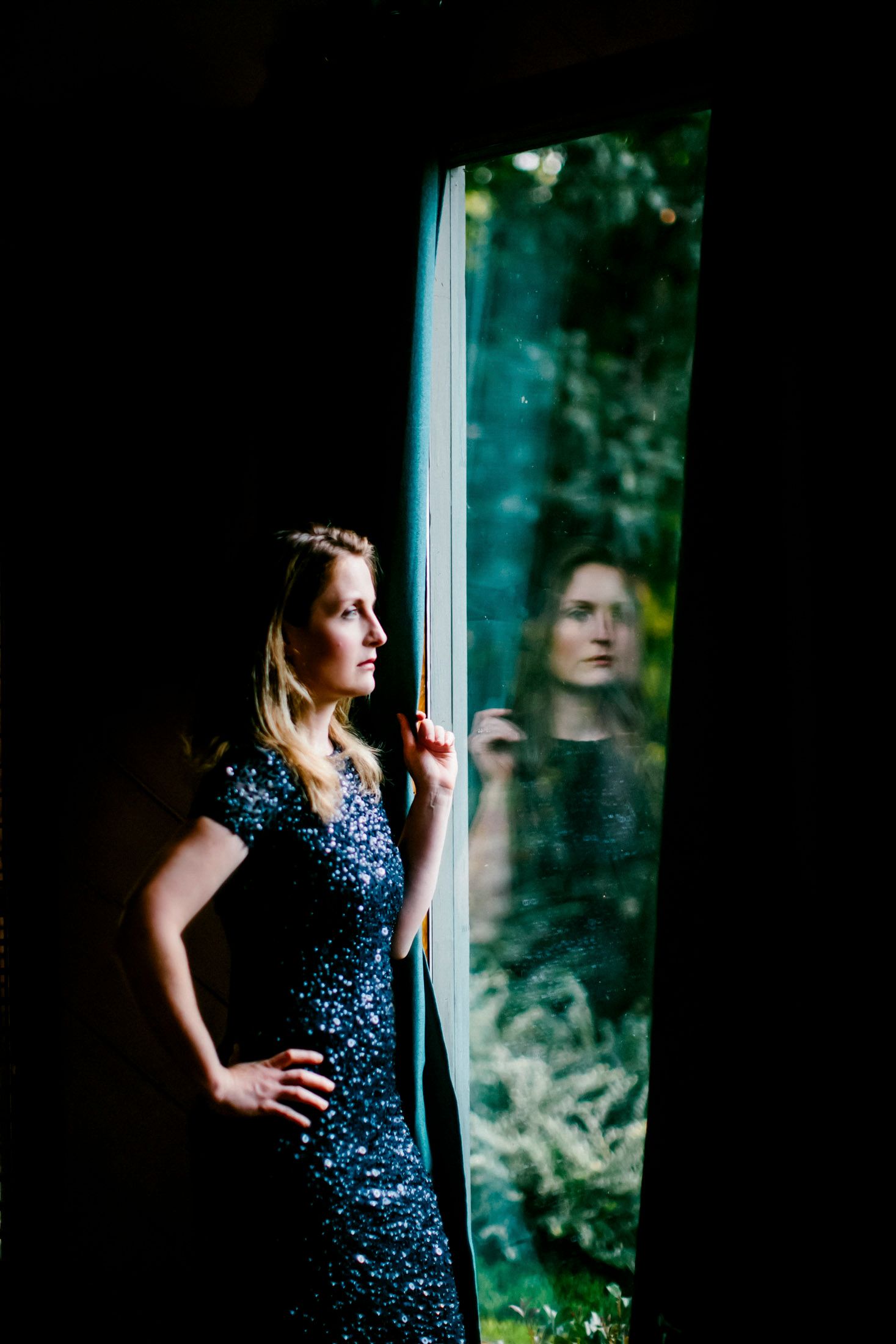 Woman in a navy sequin dress standing beside a dark window, with soft light and a reflection in the glass
