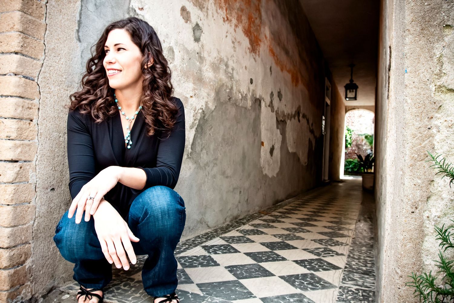 Woman with long curly hair crouching in a historic alley with a black and white checkered floor