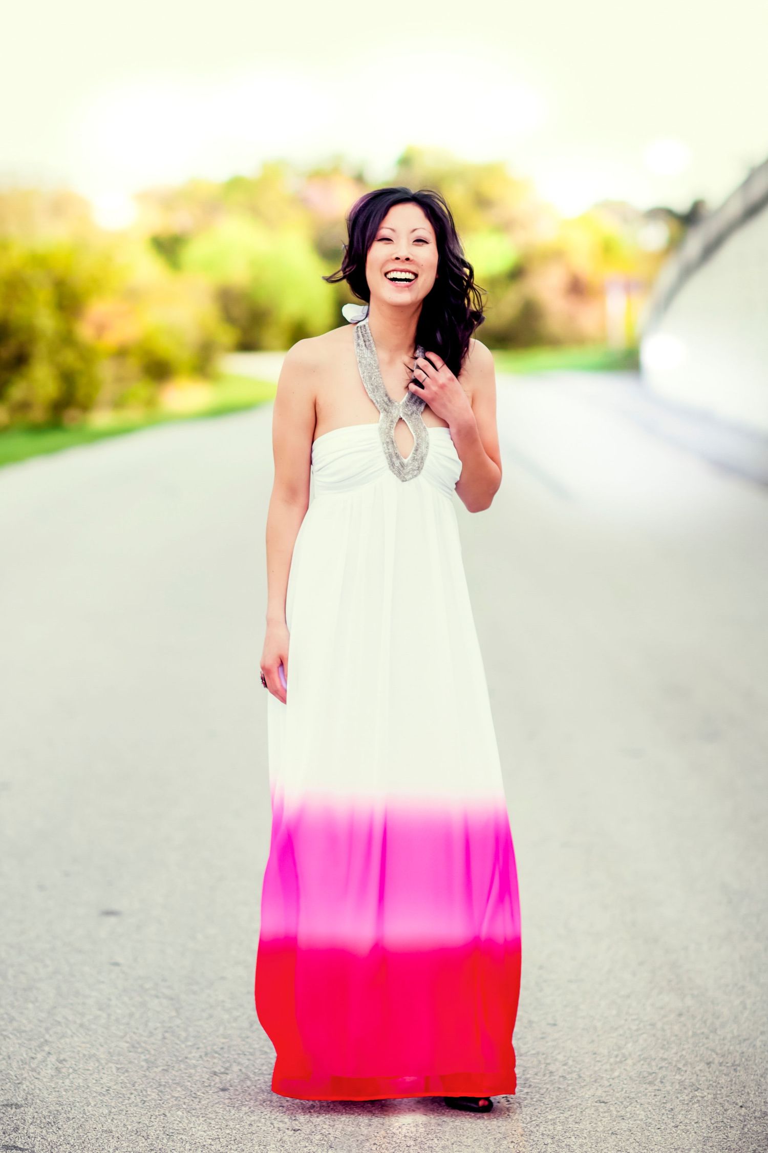 Laughing woman in a long dip-dyed pink and red maxi dress with a statement necklace, standing on a country road