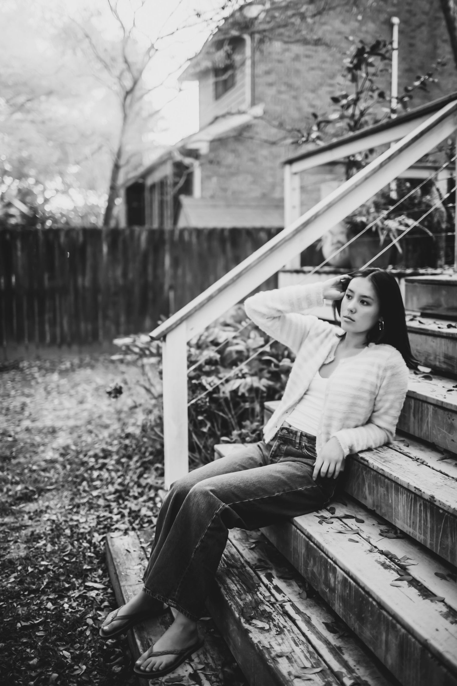 Black and white portrait of a young woman in jeans and a cardigan sitting on weathered wooden steps outdoors