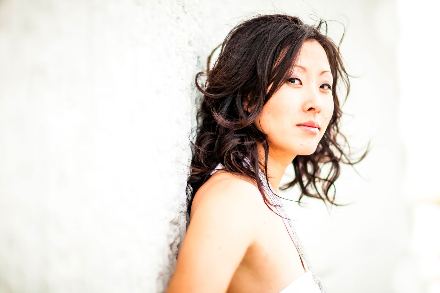 Bright, airy portrait of a woman with long dark wavy hair leaning against a white wall in soft light