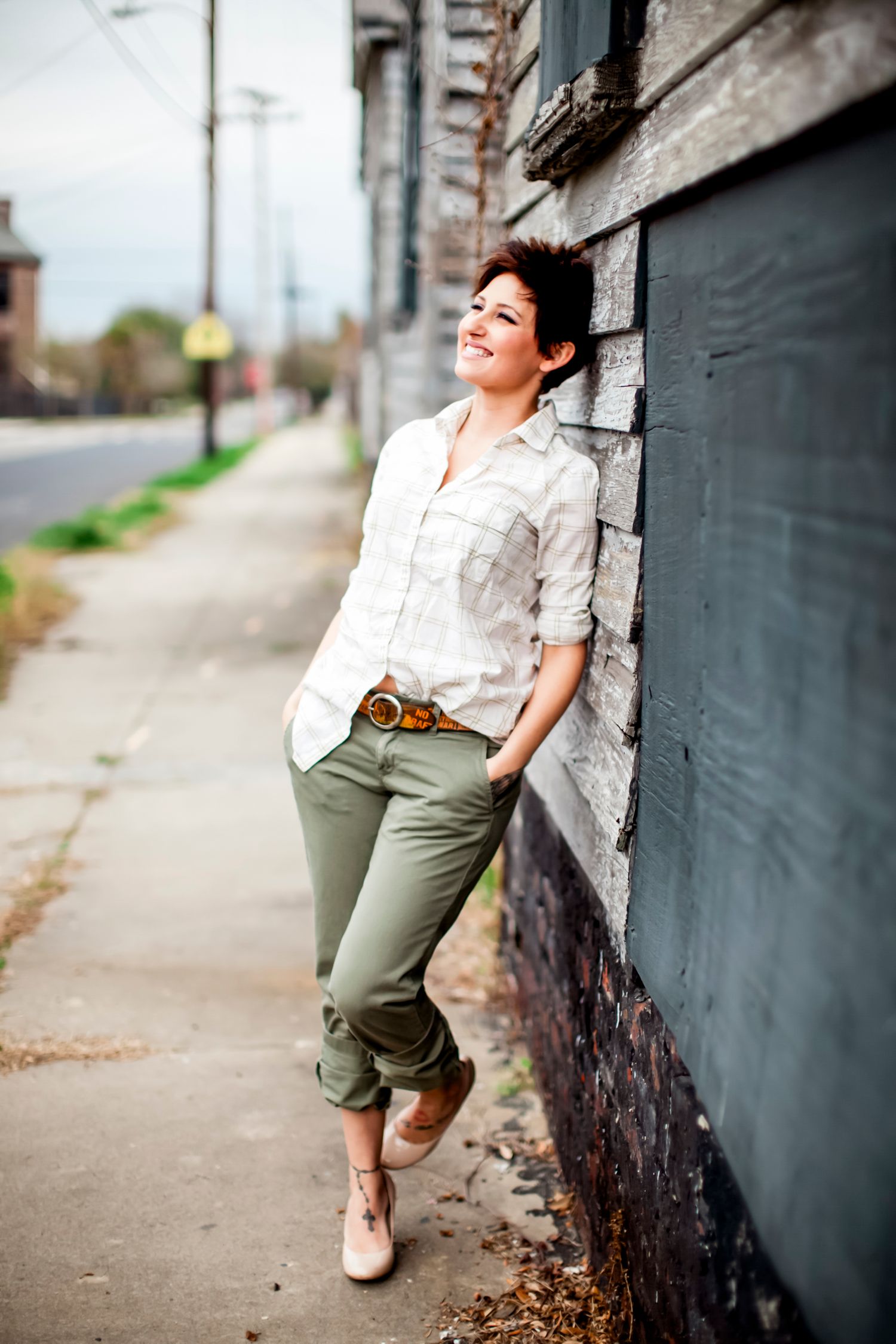 Woman in a white button-up shirt and olive pants leaning against a weathered building on a quiet street, smiling upward
