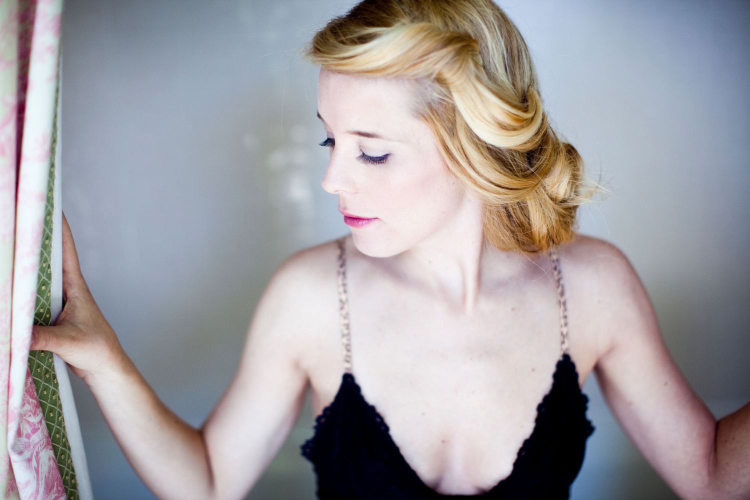 Woman with soft blonde vintage waves and a black lace camisole looking downward, one hand resting on a curtain