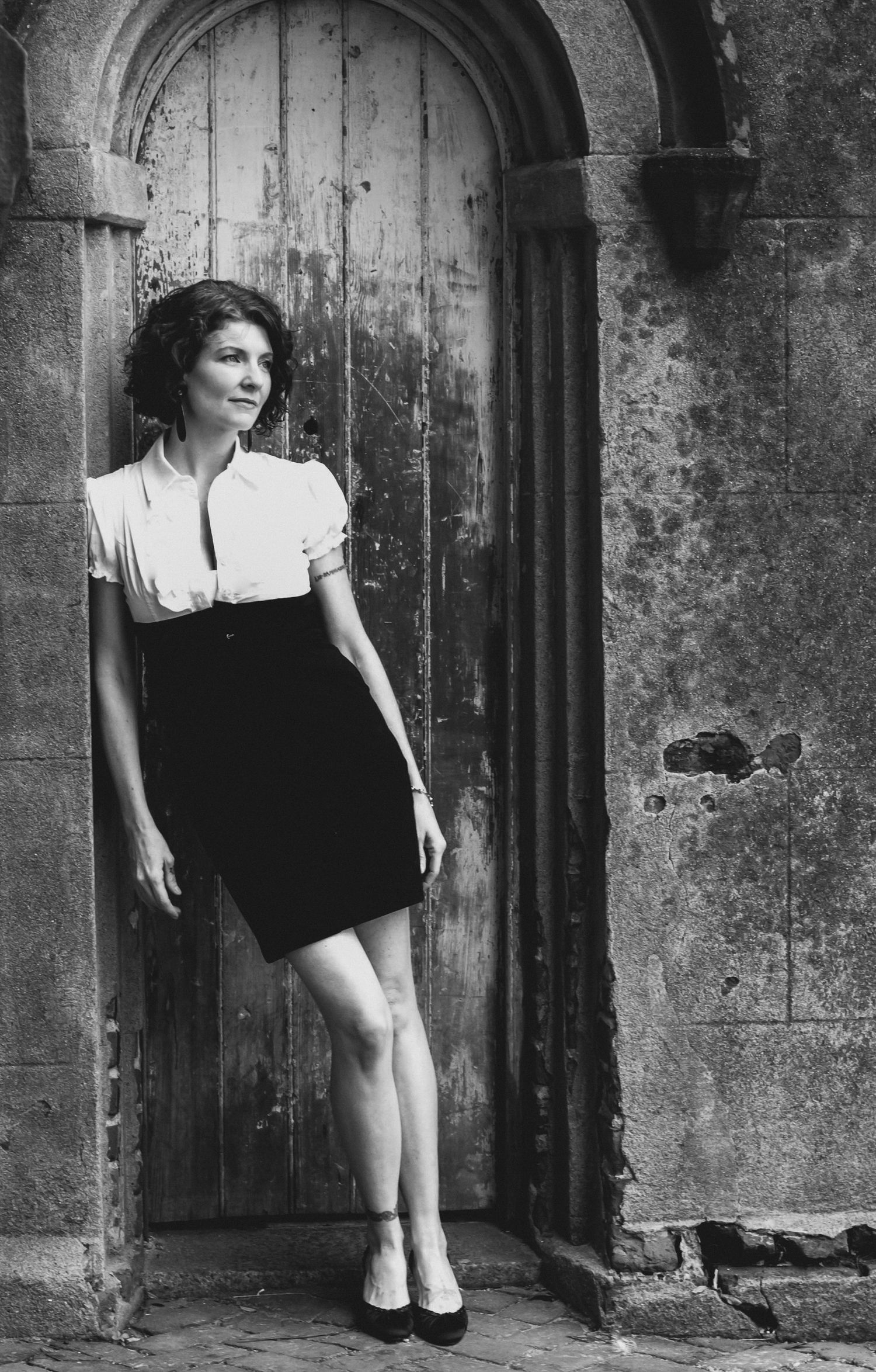 Black and white portrait of a woman in a black skirt and white collared top leaning in a weathered arched wooden doorway