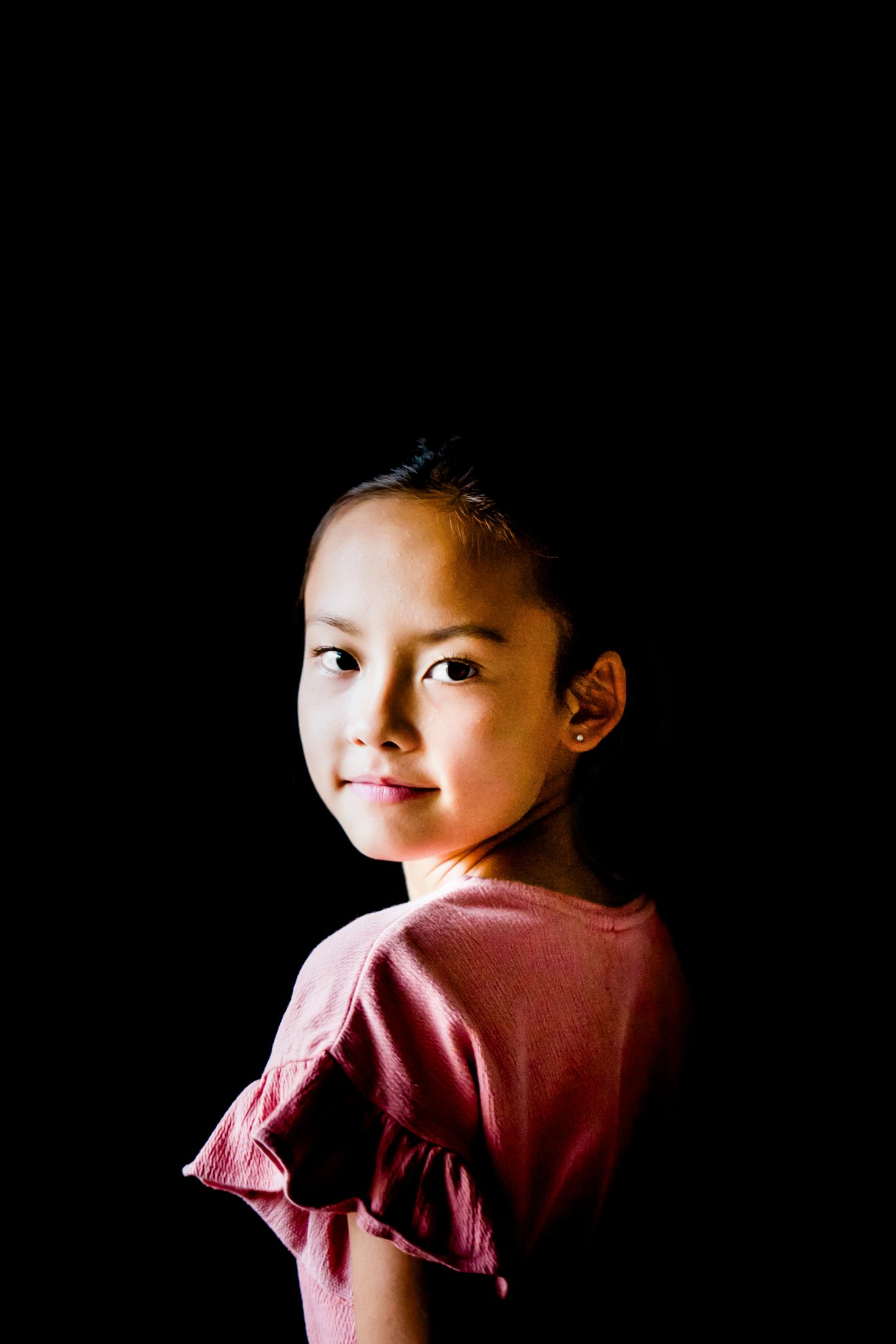 Portrait of a young girl in a dusty rose top looking over her shoulder, softly lit against a deep black background