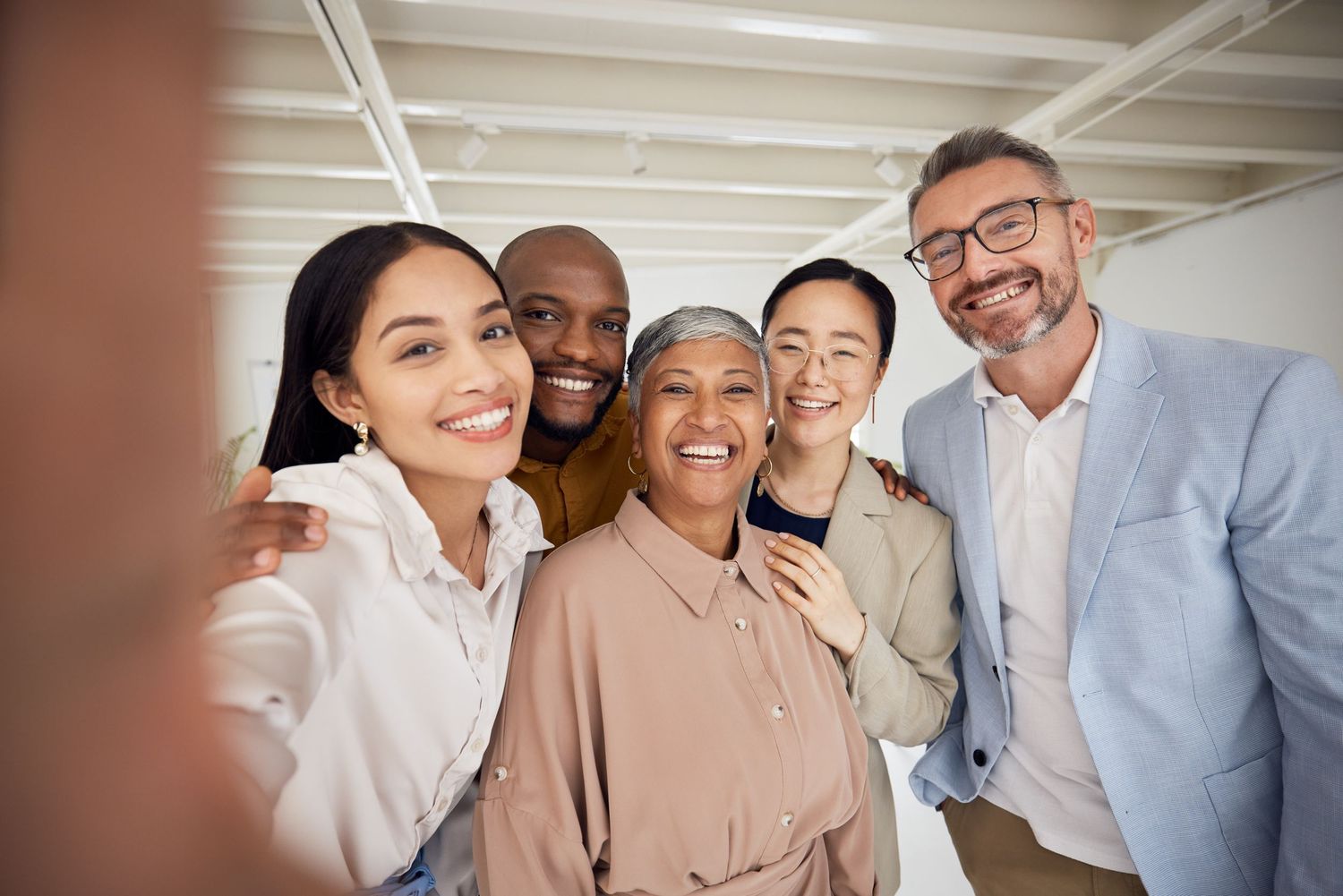 Diverse group of five smiling colleagues taking a selfie together in a bright office setting.