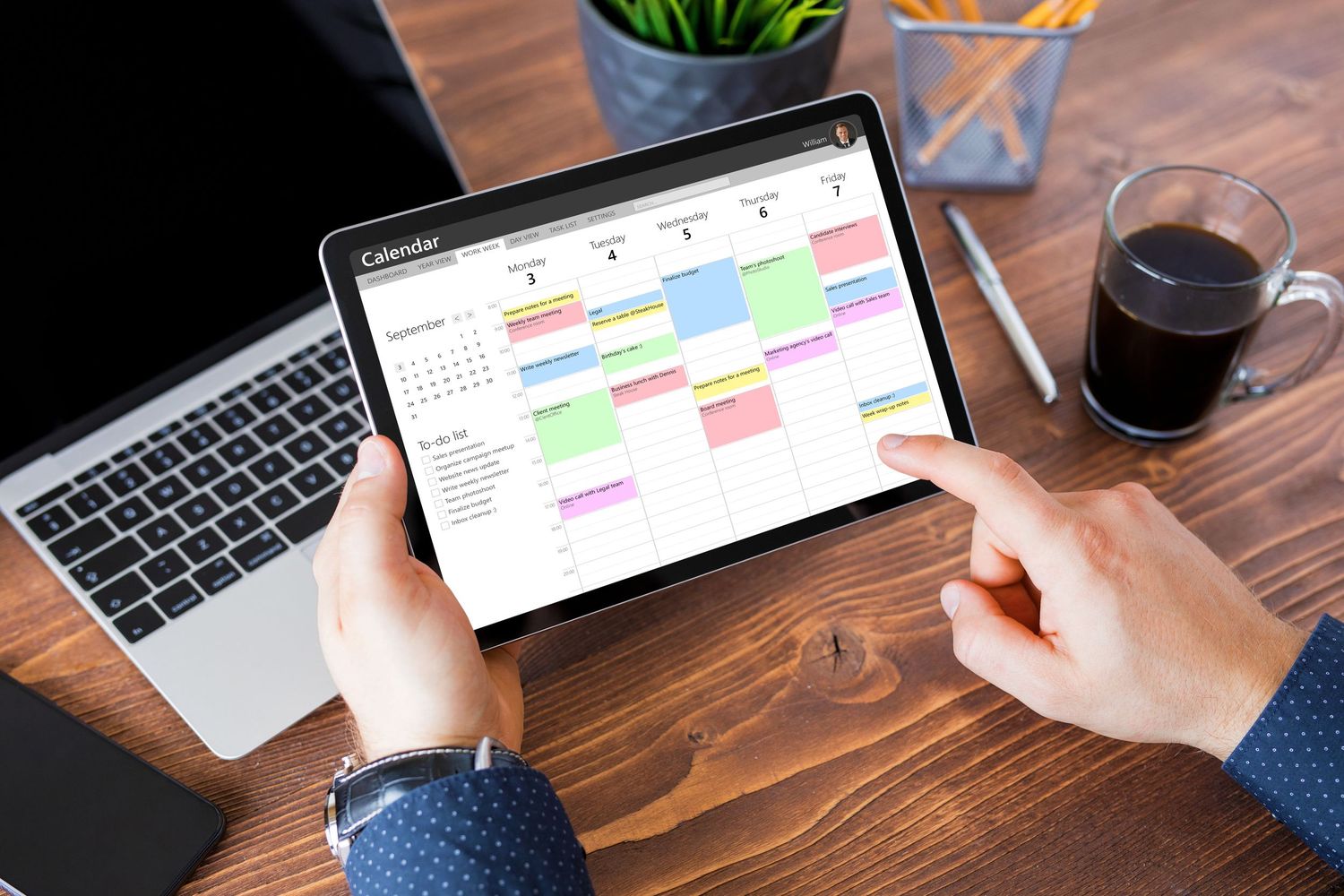 Businessman using tablet to view colorful digital calendar app on wooden desk with laptop and coffee.