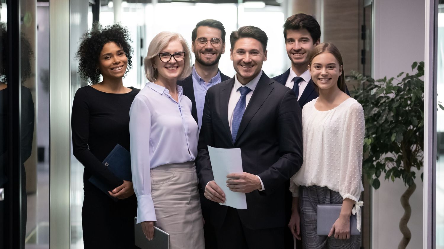Diverse group of six smiling business professionals posing together in a modern office hallway.