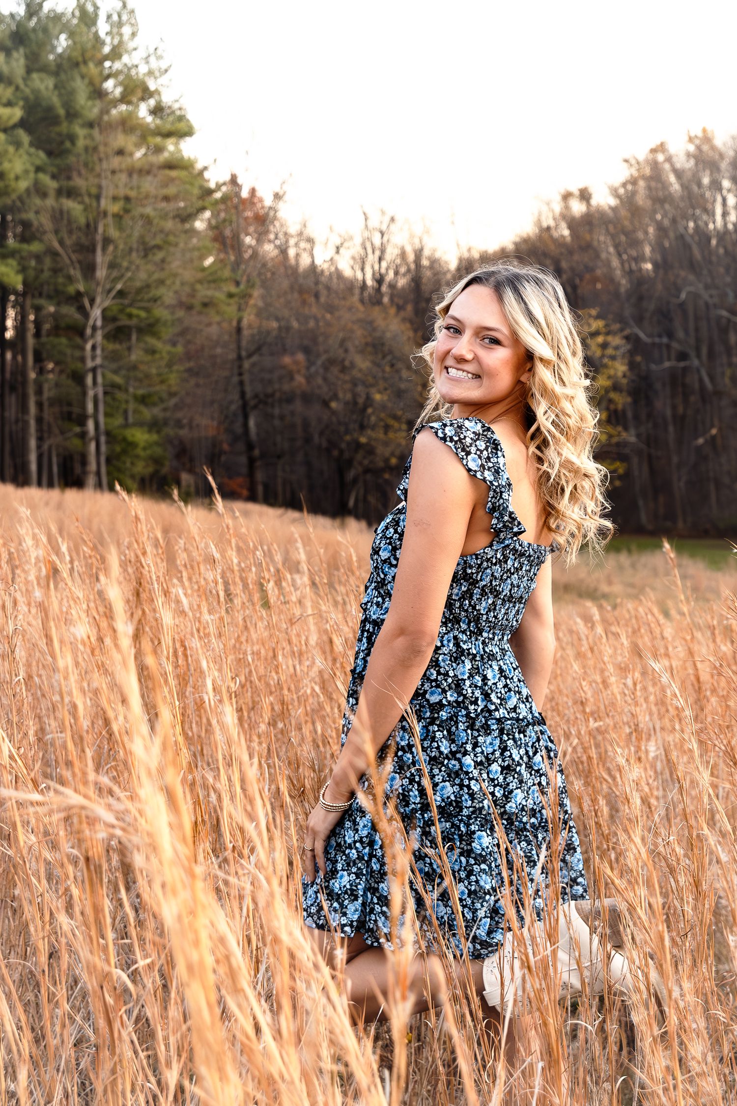 Smiling blonde woman in floral dress standing in golden field with trees in background.