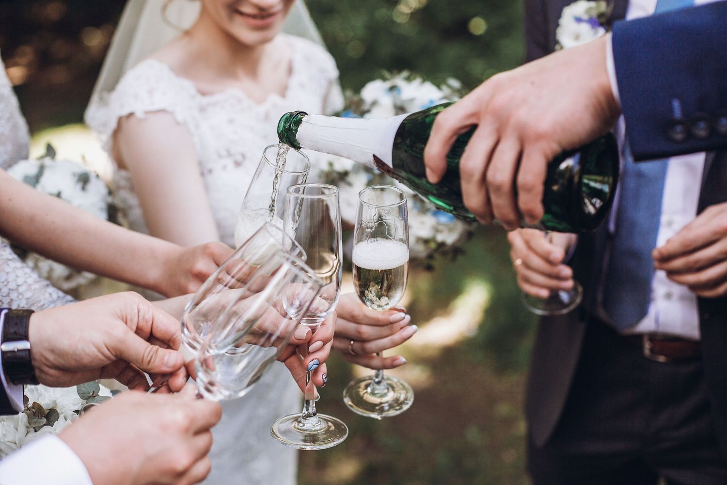 Champagne being poured into glasses at a wedding celebration with bride and guests outdoors.