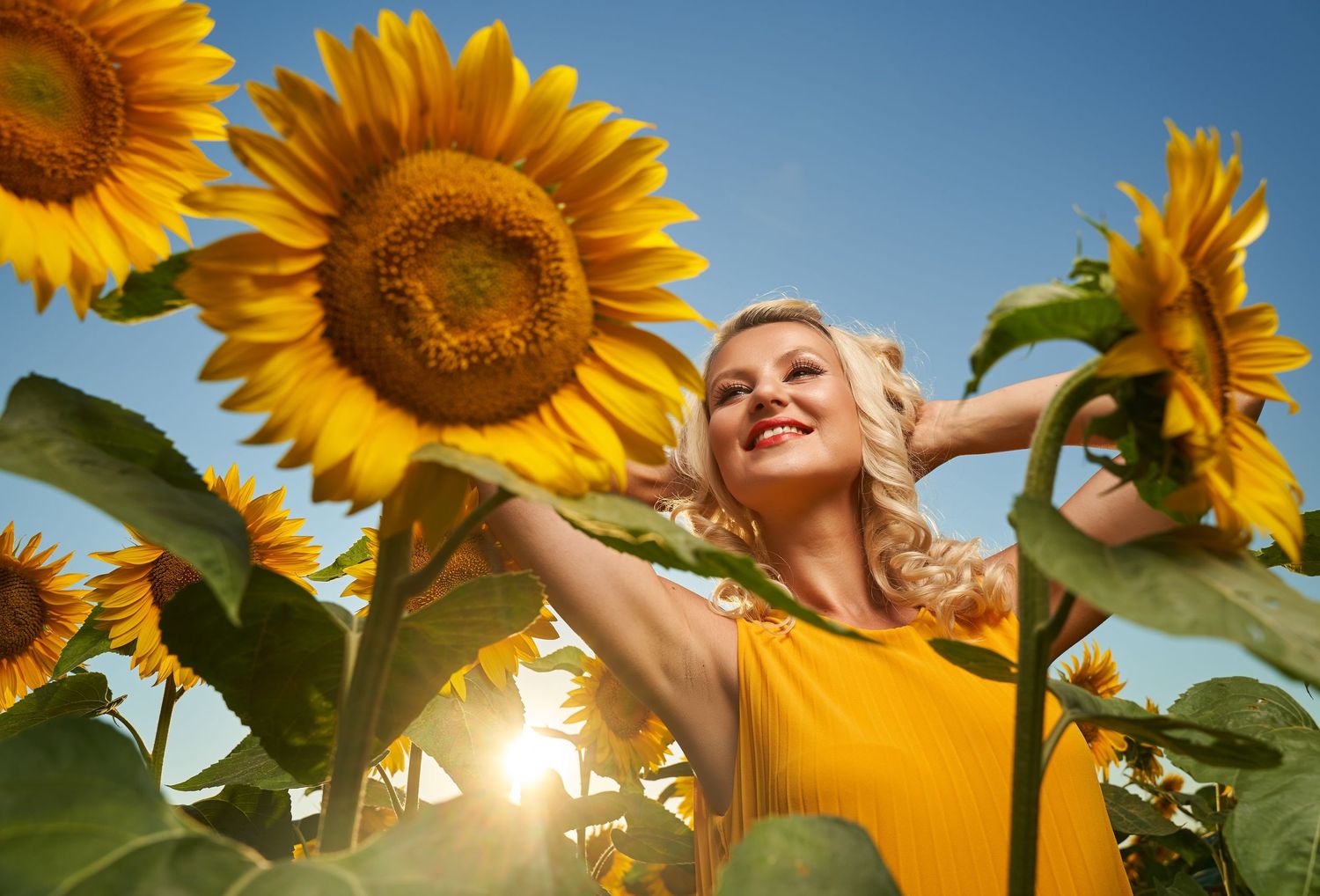 Smiling blonde woman in yellow dress surrounded by vibrant sunflowers in a sunny field.