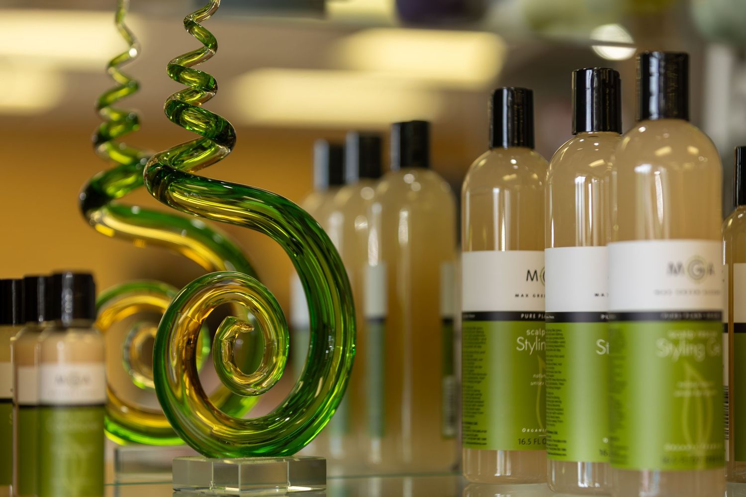 Green glass spiral sculpture displayed beside rows of M-branded lotion and shampoo bottles on a spa retail shelf.