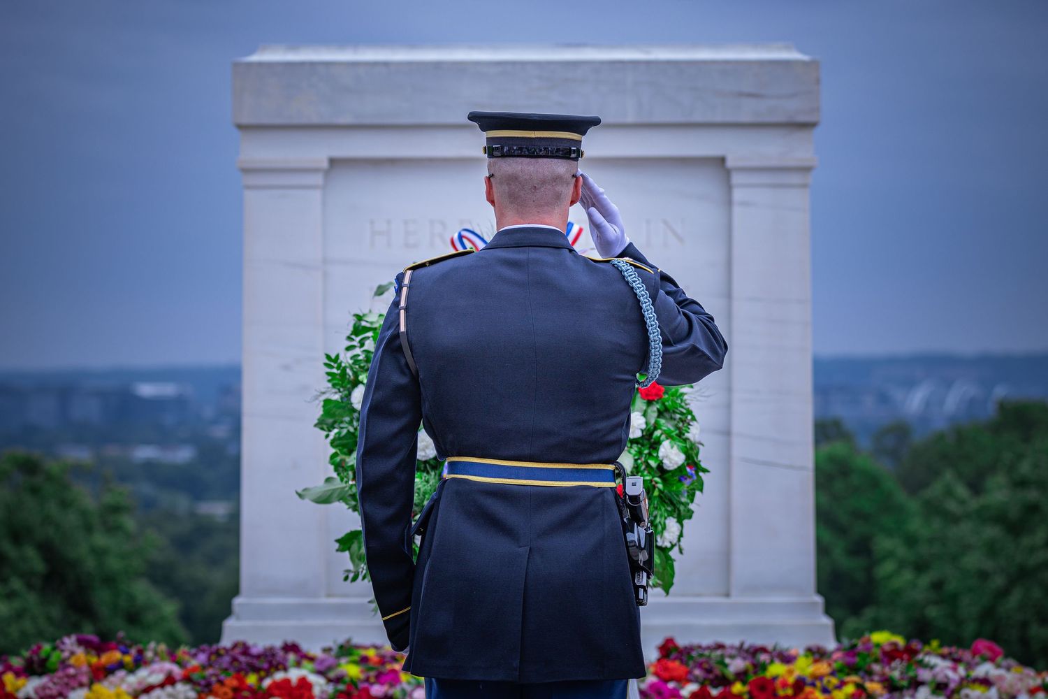 A military officer in dress uniform salutes at a memorial monument adorned with floral wreaths at dusk.