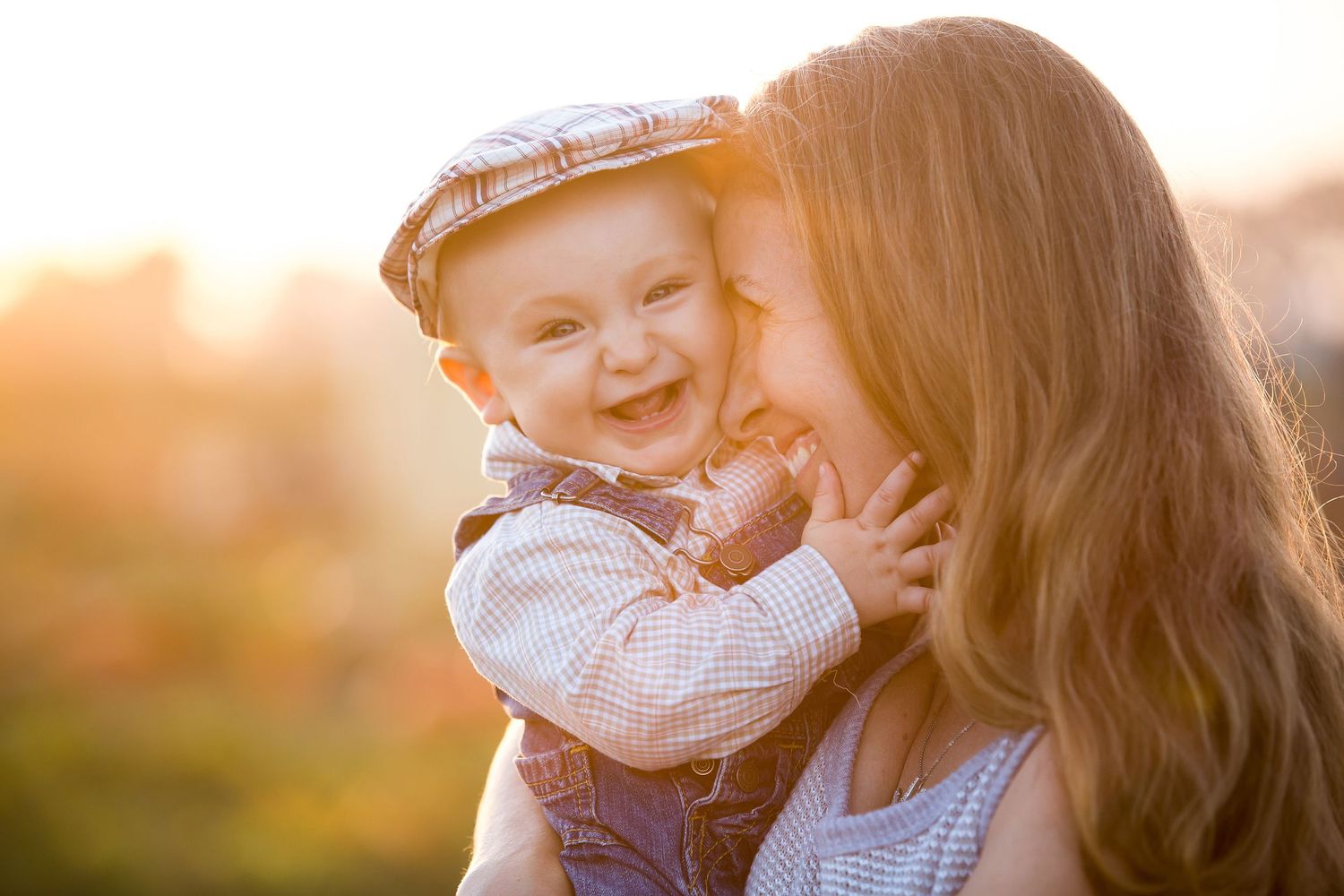 A joyful baby in a plaid outfit laughs while being held by a smiling woman in warm golden sunlight.