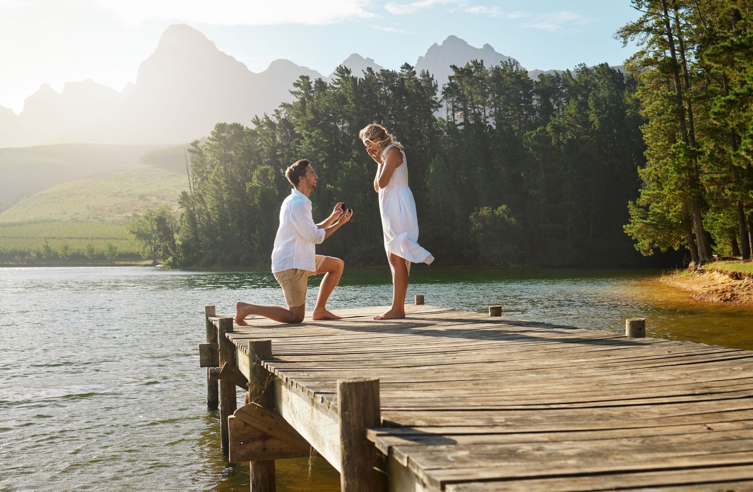 Man proposing to woman on a wooden dock by a mountain lake surrounded by lush trees in golden sunlight.