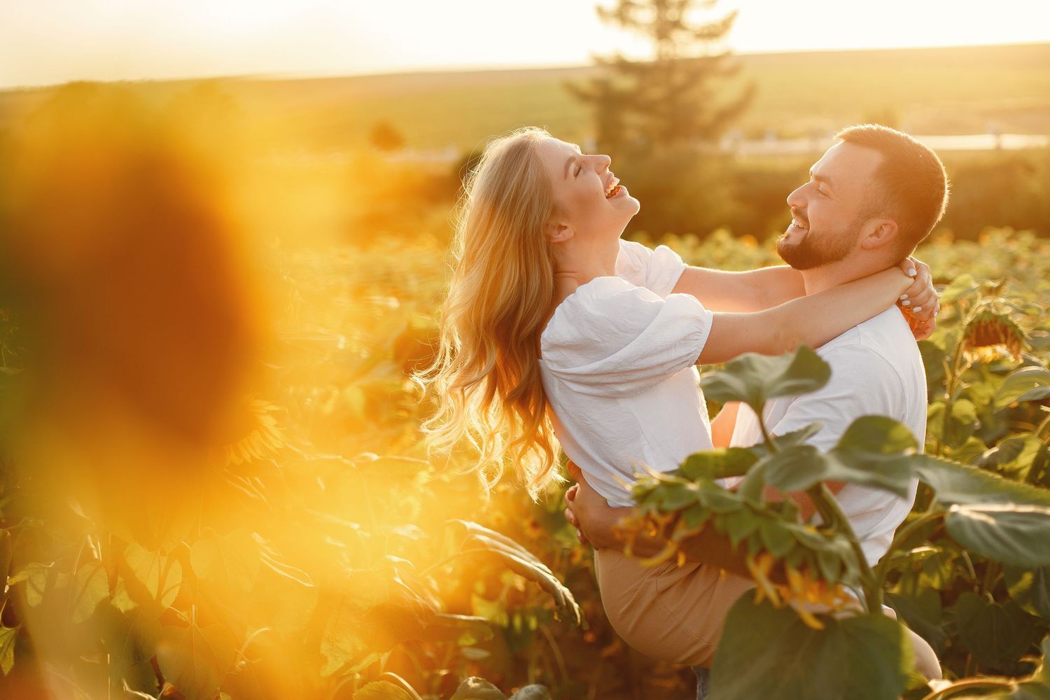 Couple embracing and laughing joyfully in a sunflower field bathed in warm golden sunset light.